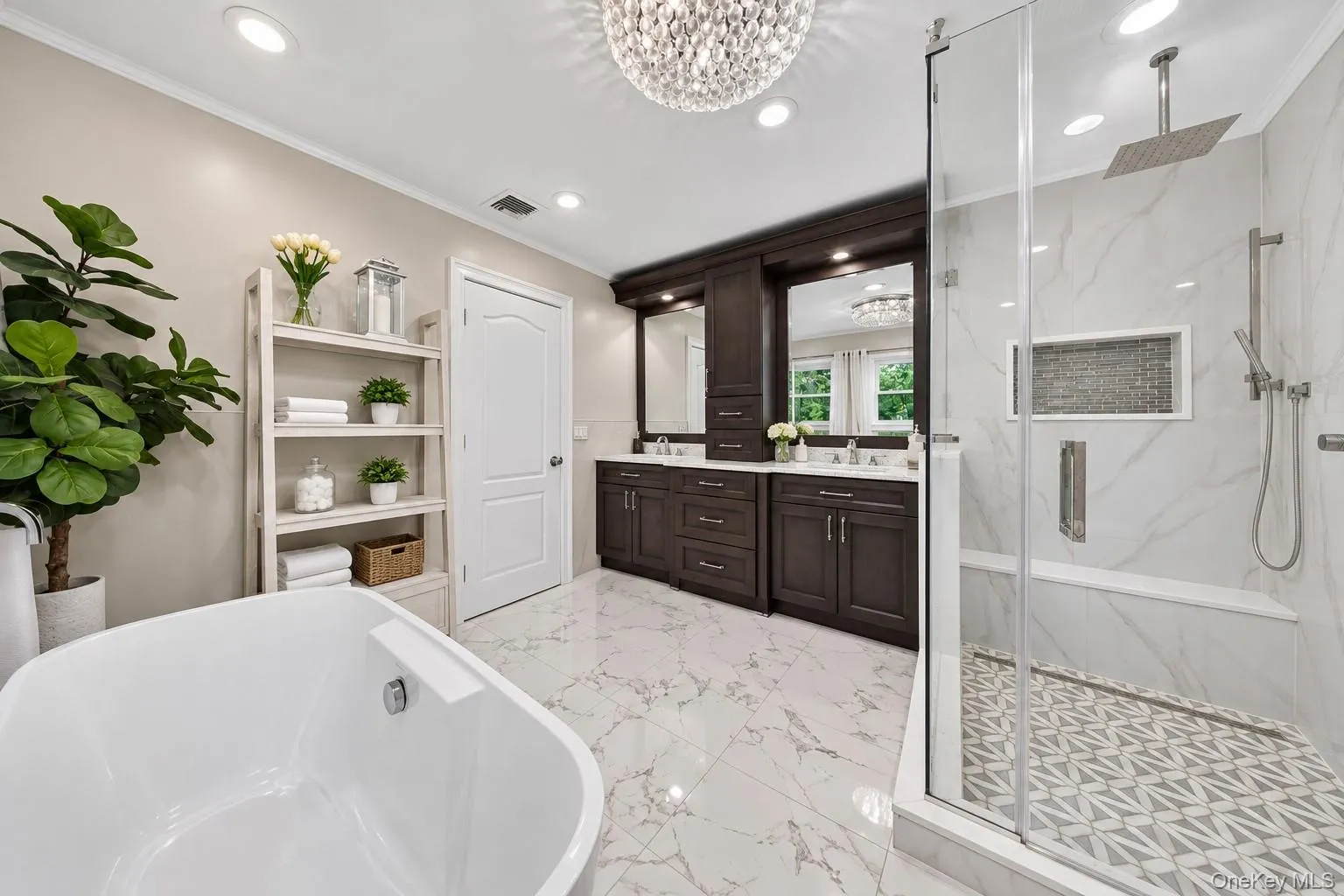 Freestanding soaking tub, dual vanity with dark wood cabinetry, glass-enclosed shower with tiled bench, rain shower head, and marble-look tile flooring Freestanding soaking tub, dual vanity with dark wood cabinetry, glass-enclosed shower with tiled bench, rain shower head, and marble-look tile flooring