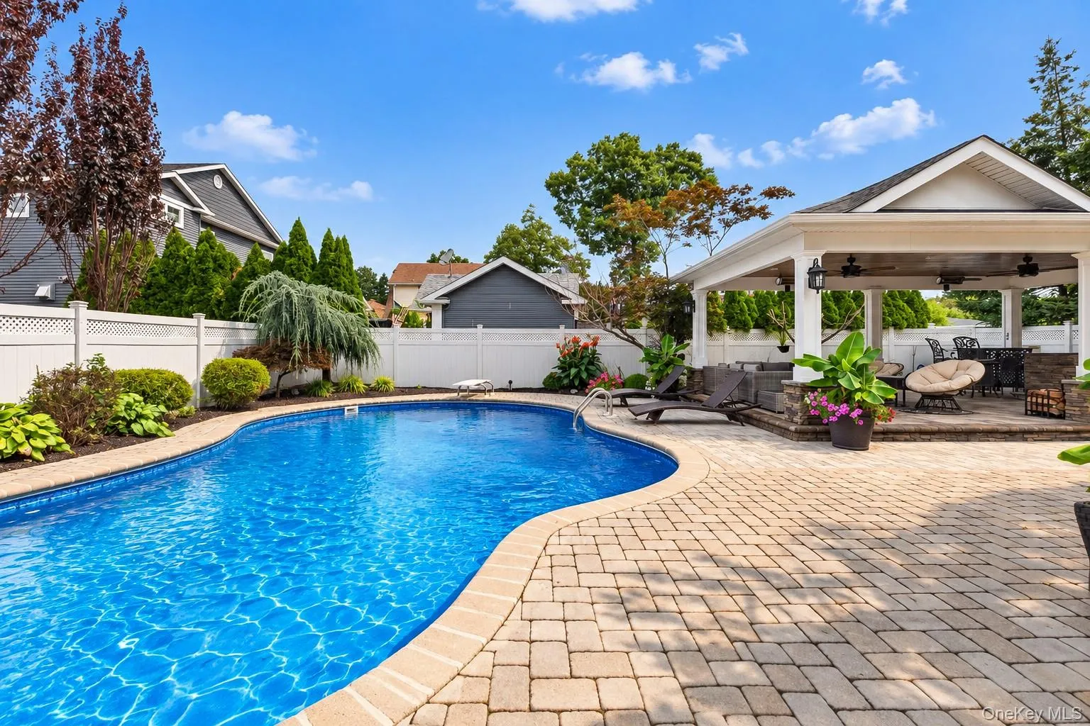 Curved inground pool with paver surround, a covered outdoor living area featuring a built-in bar, and a white lattice fence Curved inground pool with paver surround, a covered outdoor living area featuring a built-in bar, and a white lattice fence