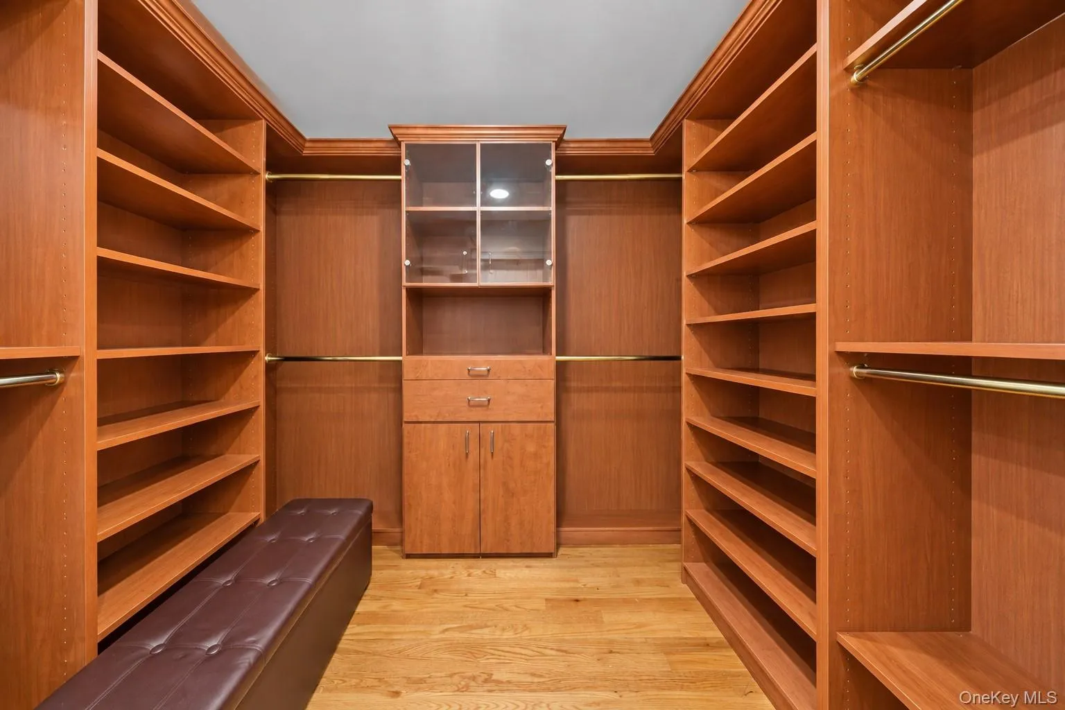Custom closet system featuring built-in shelving, hanging rods, central cabinetry with glass-front display, and wood-finish flooring Custom closet system featuring built-in shelving, hanging rods, central cabinetry with glass-front display, and wood-finish flooring
