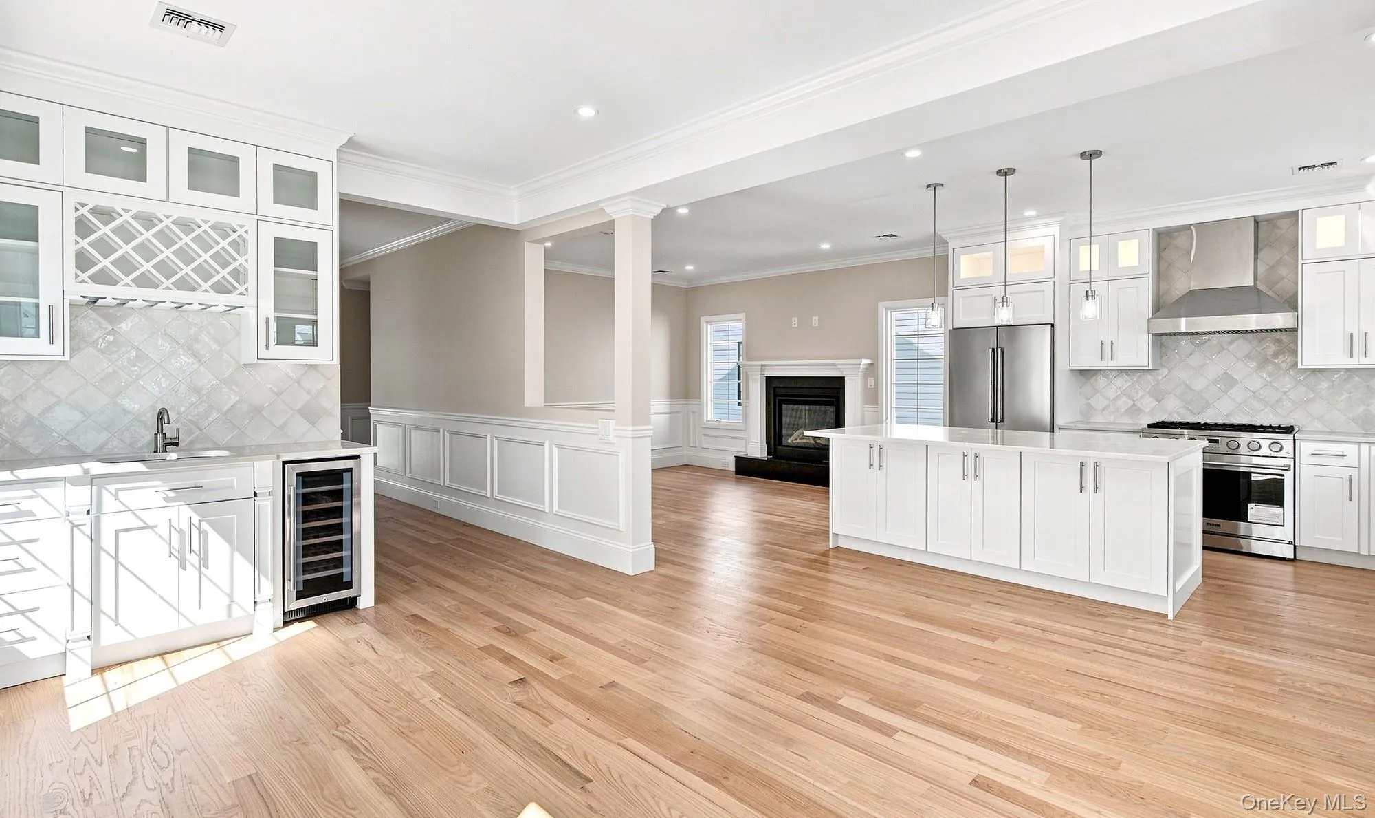 Great Room cross-view of kitchen, family room and dining. Light wood-finish flooring throughout, white cabinetry with glass-front uppers, stainless steel appliances, central kitchen island with pendant lighting, and a built-in beverage refrigerator Great Room cross-view of kitchen, family room and dining. Light wood-finish flooring throughout, white cabinetry with glass-front uppers, stainless steel appliances, central kitchen island with pendant lighting, and a built-in beverage refrigerator