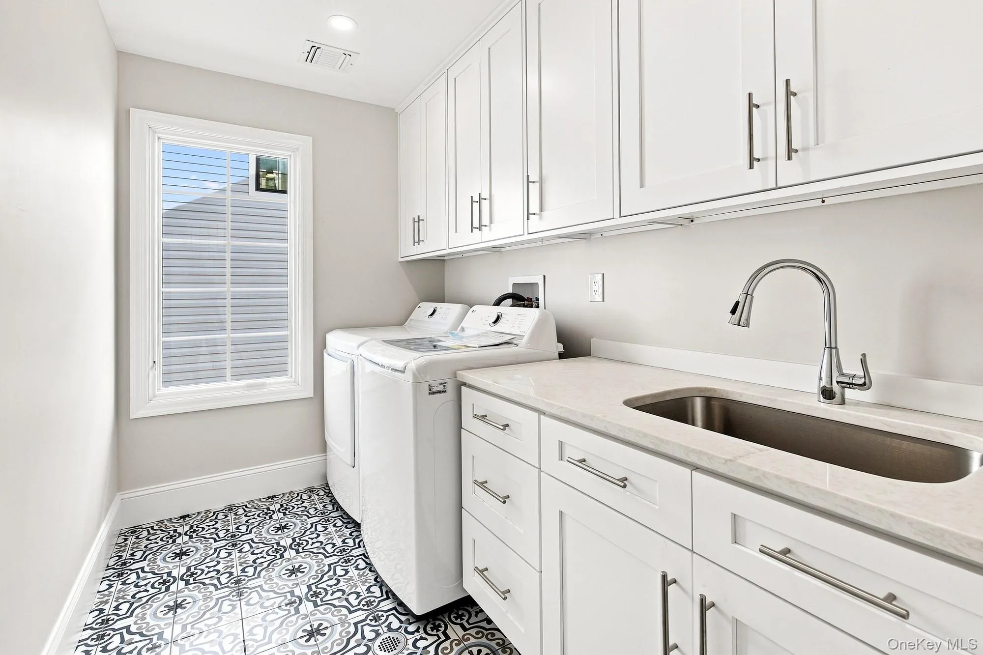 spacious Laundry Room with white shaker-style cabinetry with brushed nickel hardware, light-toned countertop, under mount stainless steel sink with gooseneck faucet, and patterned floor tile. spacious Laundry Room with white shaker-style cabinetry with brushed nickel hardware, light-toned countertop, under mount stainless steel sink with gooseneck faucet, and patterned floor tile.