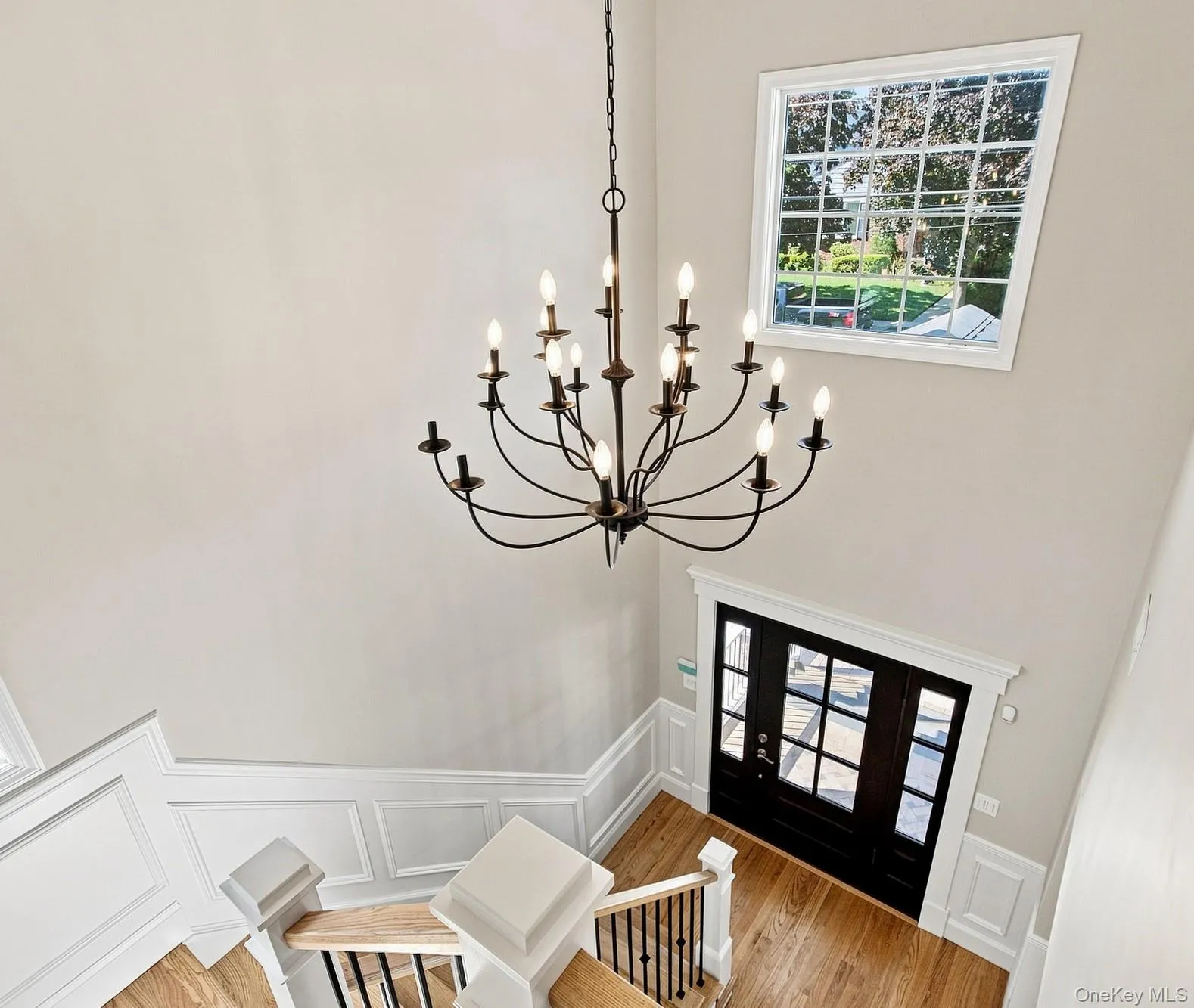 Spacious foyer featuring a multi-tier chandelier, wood-finish flooring, white panel molding, and a dark-toned entry door with multiple glass panes. Spacious foyer featuring a multi-tier chandelier, wood-finish flooring, white panel molding, and a dark-toned entry door with multiple glass panes.