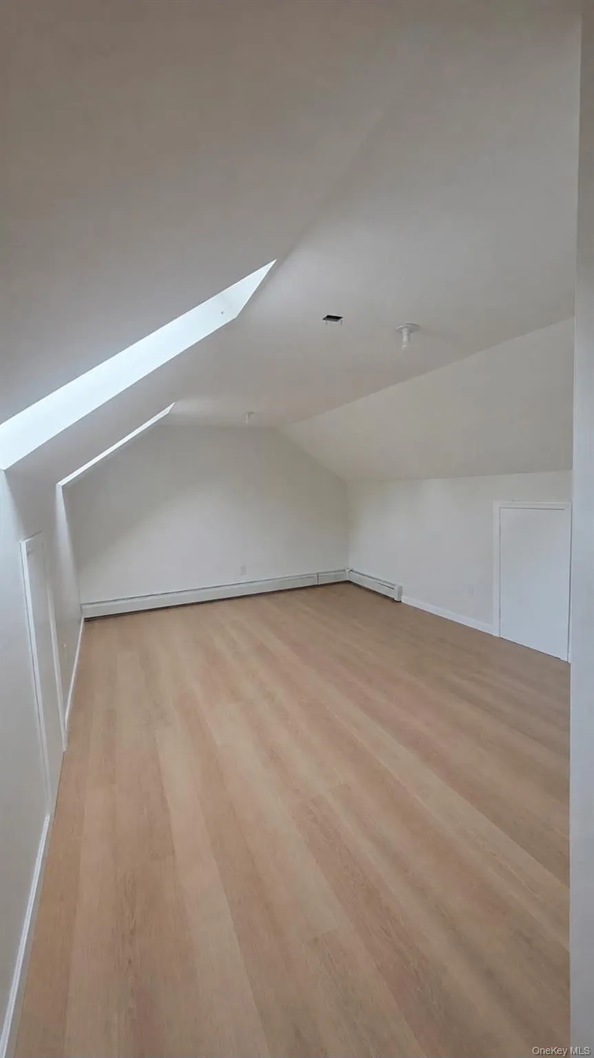 Finished attic with vaulted ceiling, skylight, wood-finish flooring, white walls, and baseboard heating Finished attic with vaulted ceiling, skylight, wood-finish flooring, white walls, and baseboard heating