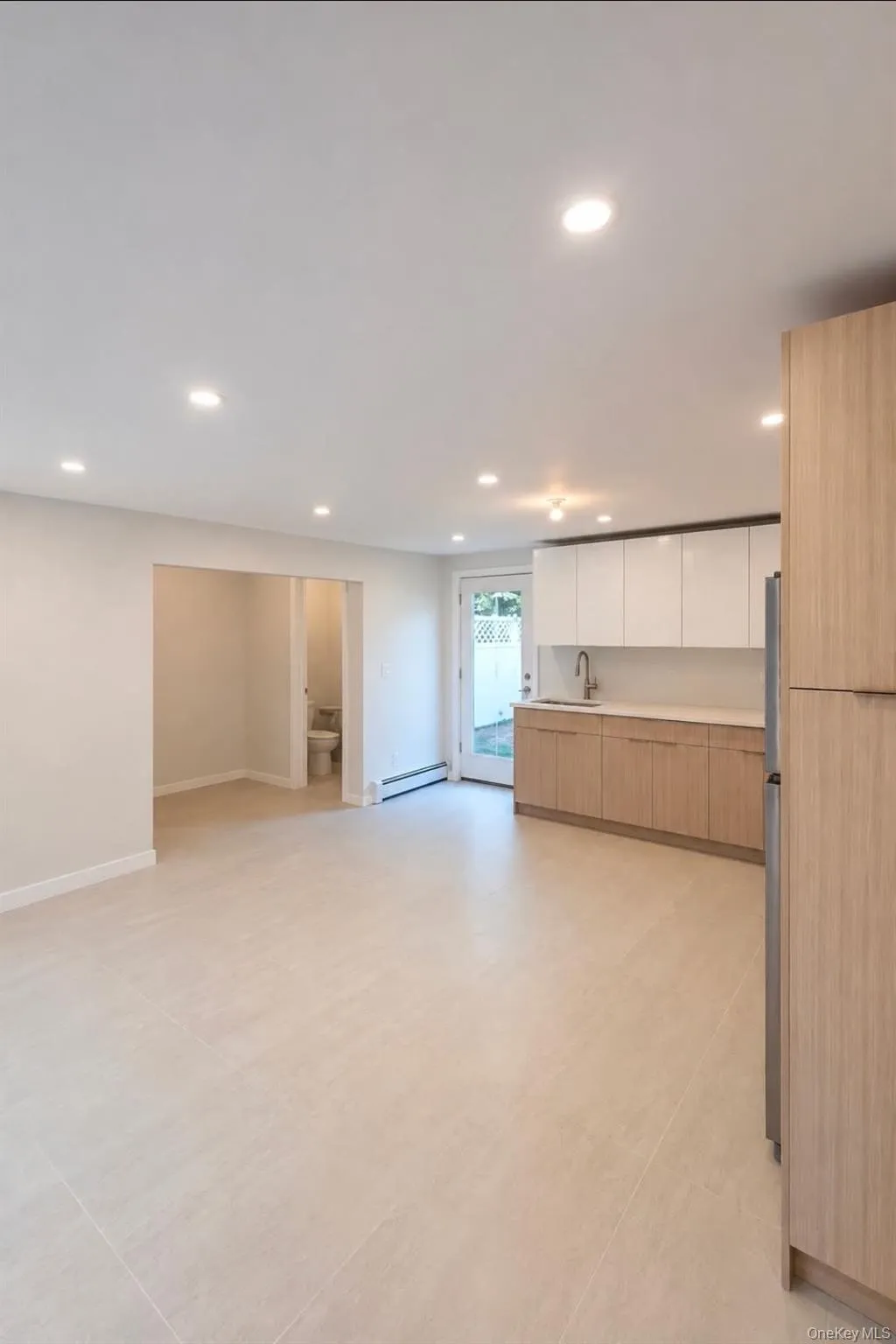 Recessed lighting throughout, light-toned tile flooring, contemporary kitchen cabinetry with a light wood finish and white upper cabinets, stainless steel refrigerator, and a door providing exterior access Recessed lighting throughout, light-toned tile flooring, contemporary kitchen cabinetry with a light wood finish and white upper cabinets, stainless steel refrigerator, and a door providing exterior access