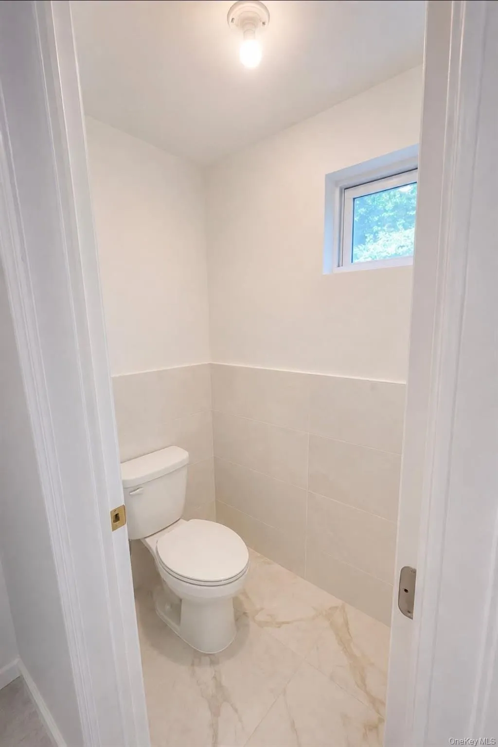 White ceramic toilet, half-wall tiled wainscoting, marble-look tiled flooring, and a window for natural light White ceramic toilet, half-wall tiled wainscoting, marble-look tiled flooring, and a window for natural light