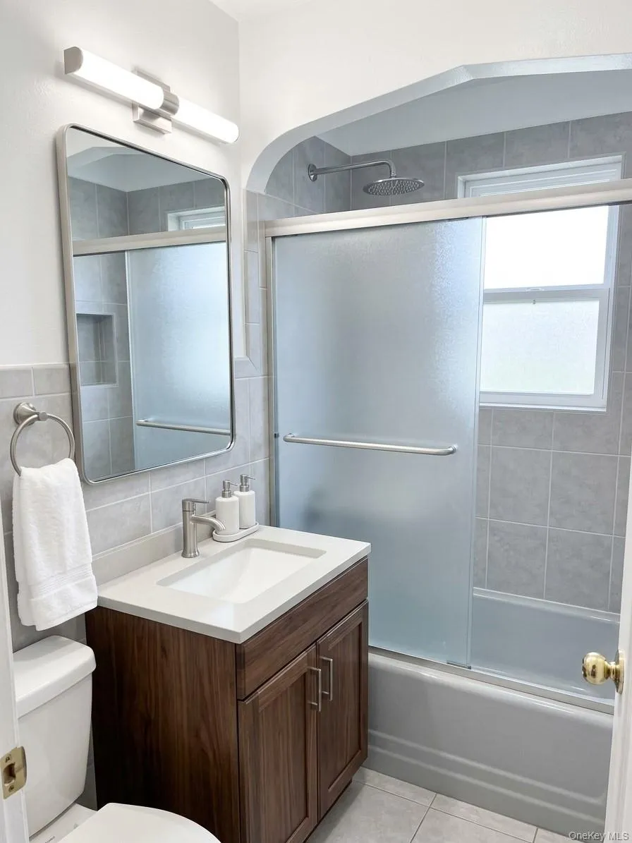 Bathroom featuring a wood-finish vanity with an integrated sink, a frosted glass shower enclosure, a rainfall showerhead, and tile flooring Bathroom featuring a wood-finish vanity with an integrated sink, a frosted glass shower enclosure, a rainfall showerhead, and tile flooring