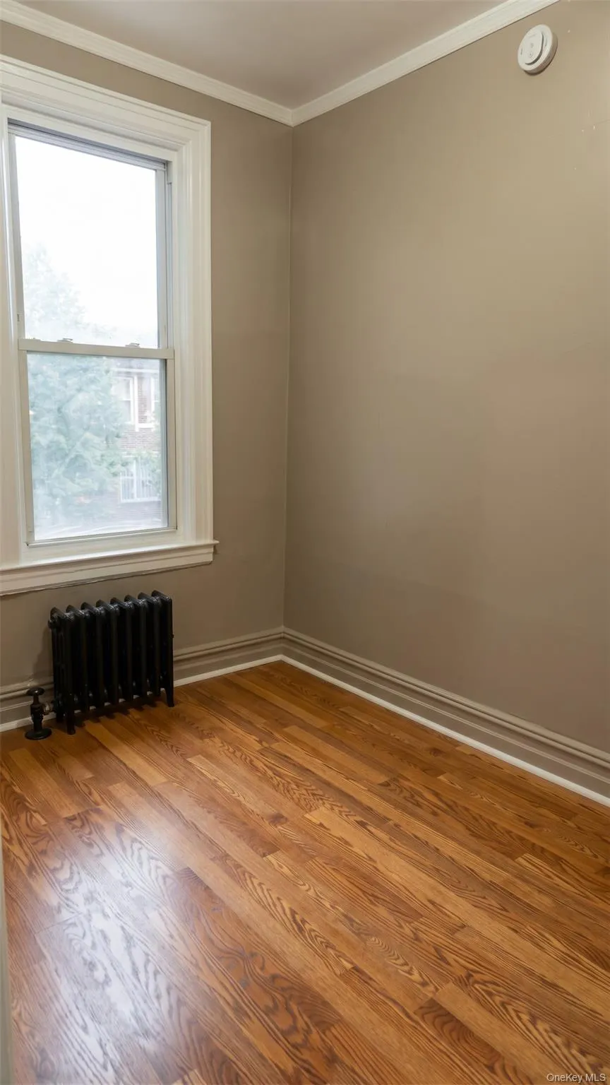 Hardwood flooring, classic window with trim, crown molding, baseboard molding, and a cast-iron radiator Hardwood flooring, classic window with trim, crown molding, baseboard molding, and a cast-iron radiator