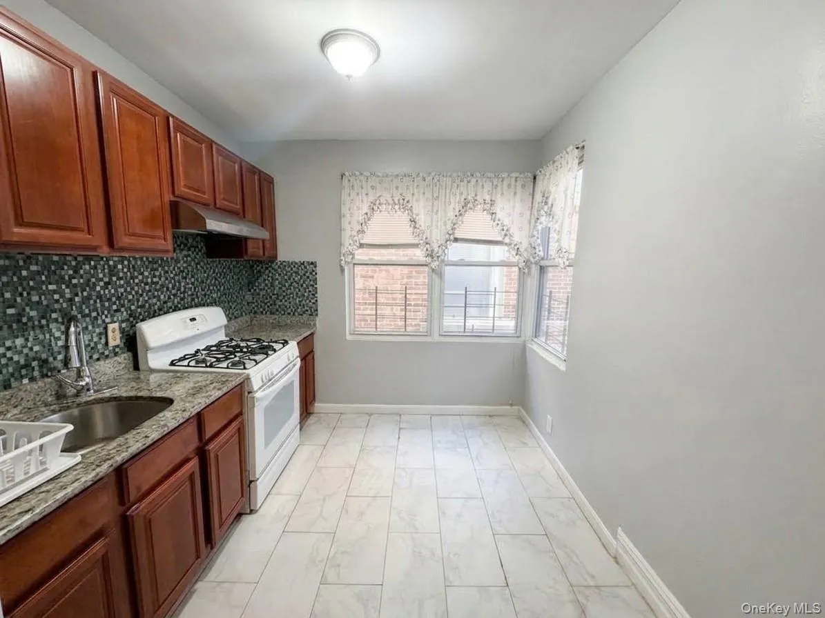 Kitchen featuring wood-finish cabinetry, patterned tile backsplash, granite-style countertops, and white tile flooring Kitchen featuring wood-finish cabinetry, patterned tile backsplash, granite-style countertops, and white tile flooring