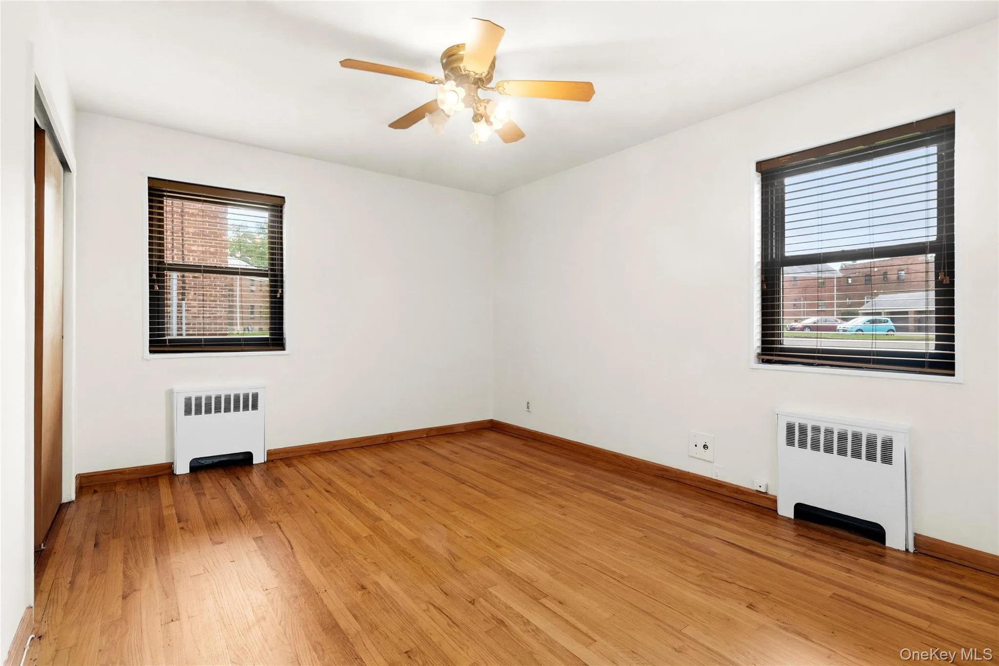 Hardwood flooring throughout, two windows with blinds, a ceiling fan with integrated lighting, and white baseboard heating units Hardwood flooring throughout, two windows with blinds, a ceiling fan with integrated lighting, and white baseboard heating units