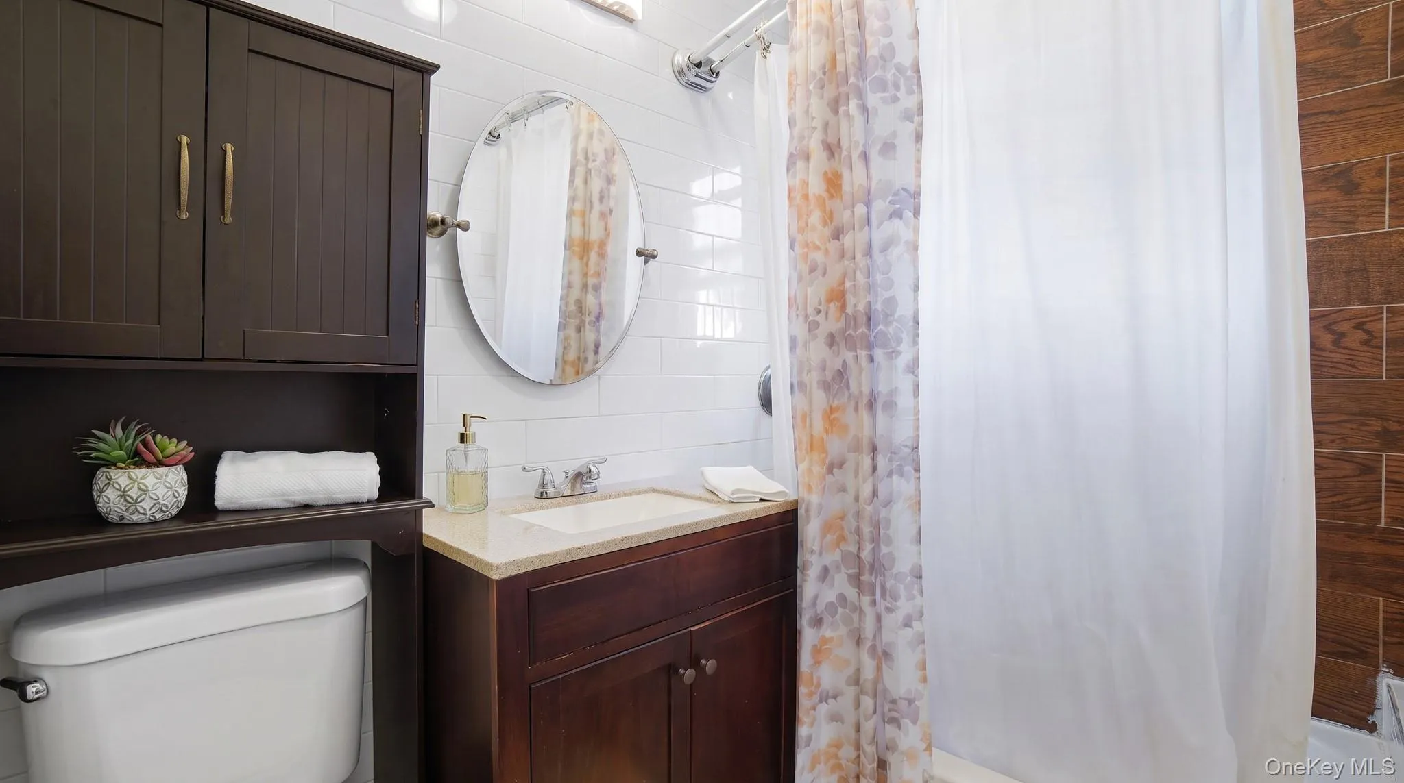 White subway tile surround, wood-finish accent wall, dark wood vanity with a light-toned countertop, and an oval mirror White subway tile surround, wood-finish accent wall, dark wood vanity with a light-toned countertop, and an oval mirror