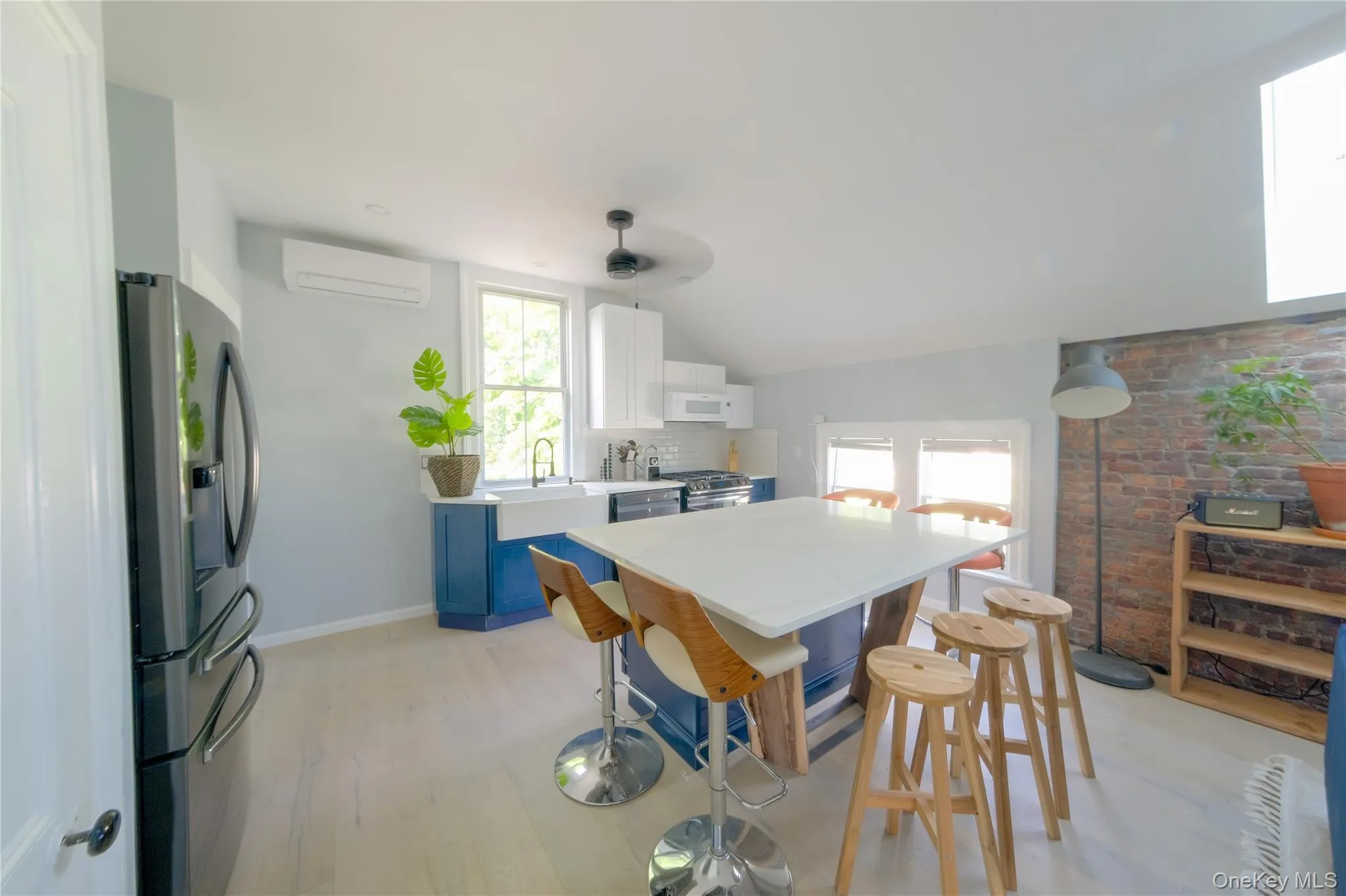 Kitchen featuring a central island with countertop, a farmhouse sink, light wood-finish flooring, exposed brick accent wall, and built-in white cabinetry Kitchen featuring a central island with countertop, a farmhouse sink, light wood-finish flooring, exposed brick accent wall, and built-in white cabinetry