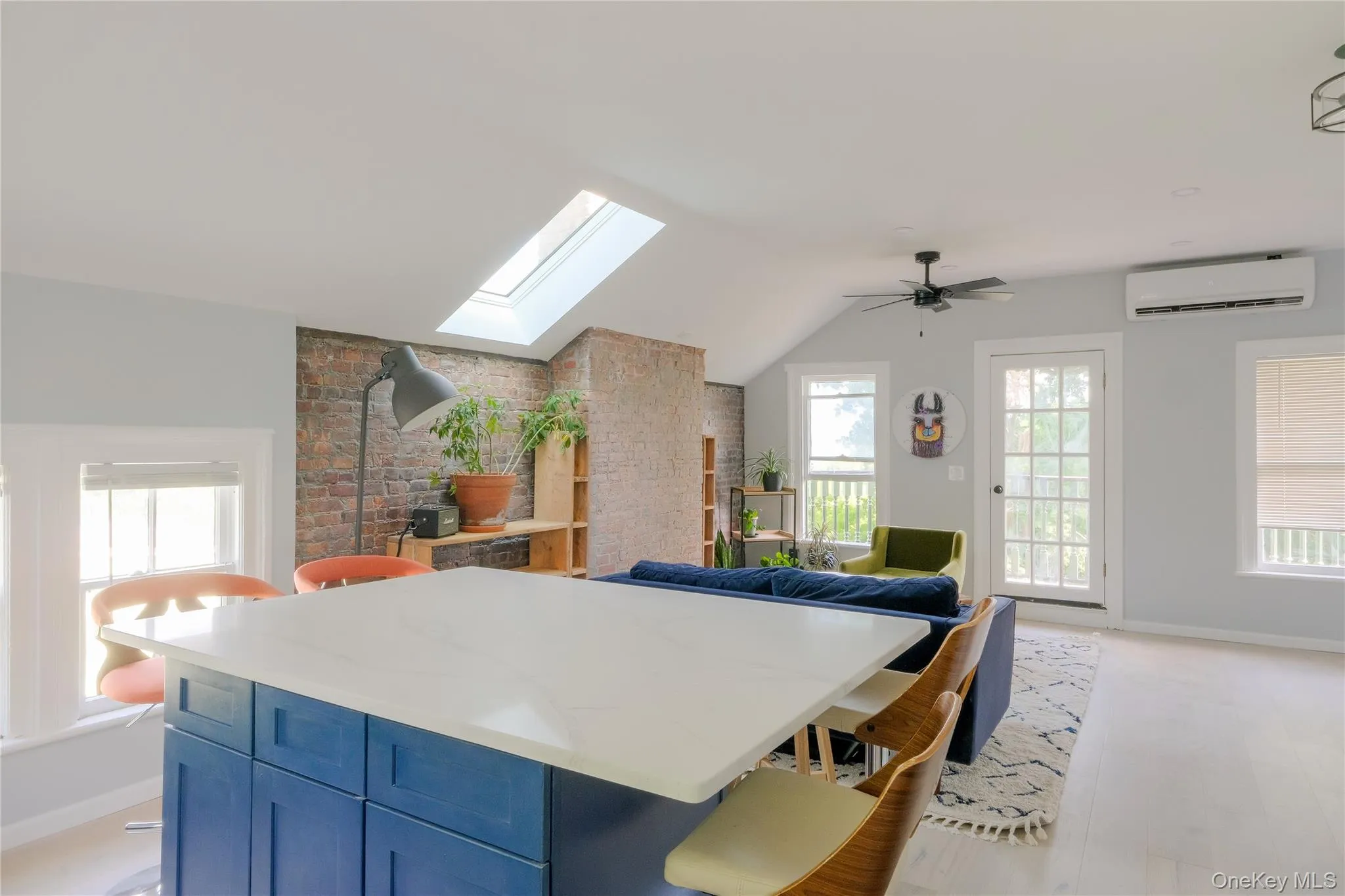 Skylight, exposed brick accent wall, white countertop island with blue cabinetry, ceiling fan, and light wood-finish flooring Skylight, exposed brick accent wall, white countertop island with blue cabinetry, ceiling fan, and light wood-finish flooring