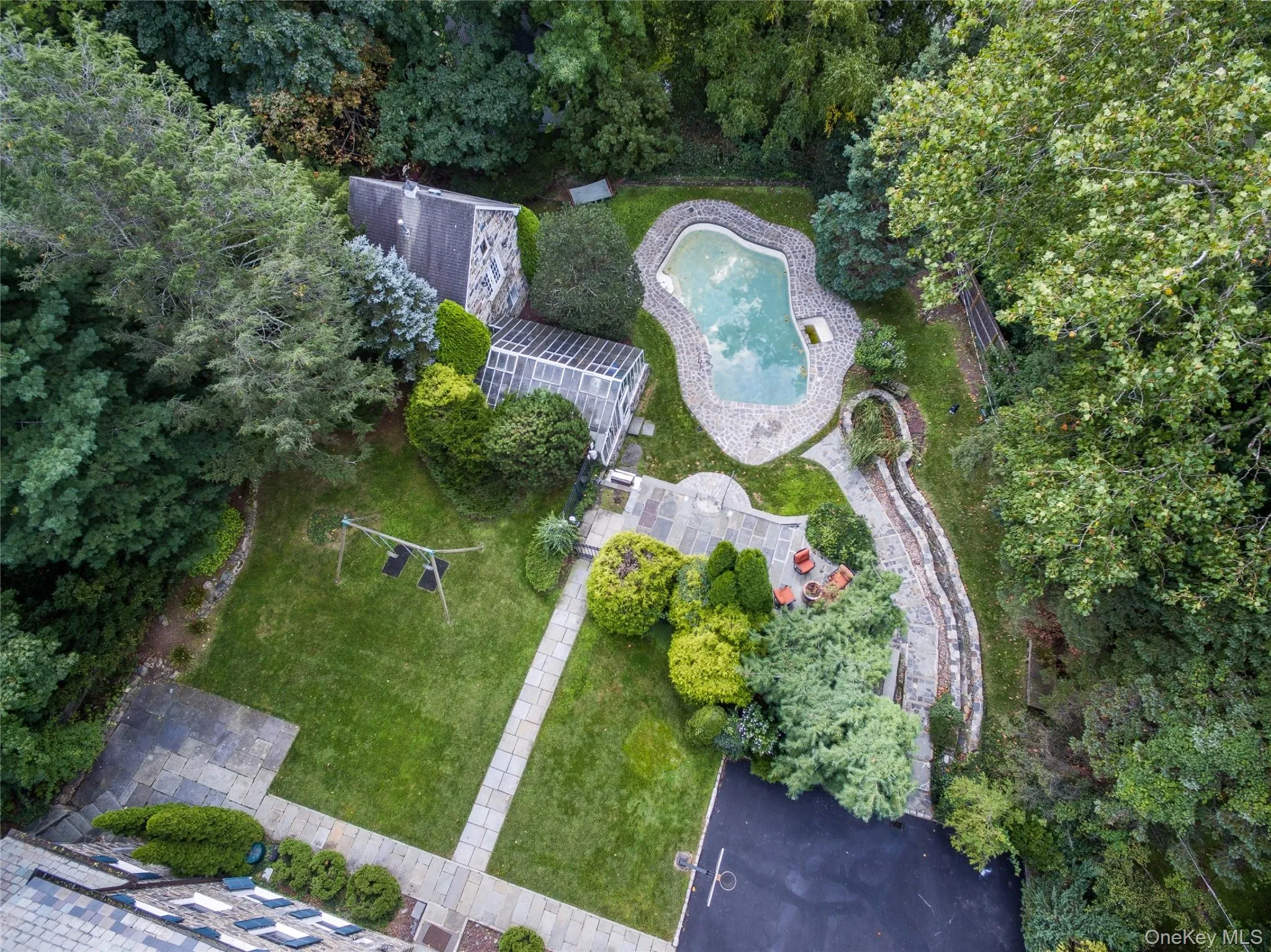 Aerial view of an expansive residential property featuring a custom-shaped swimming pool with integrated spa, manicured lawn, mature tree-lined perimeter, paved pathways, and multiple outbuildings nestled within lush landscaping Aerial view of an expansive residential property featuring a custom-shaped swimming pool with integrated spa, manicured lawn, mature tree-lined perimeter, paved pathways, and multiple outbuildings nestled within lush landscaping
