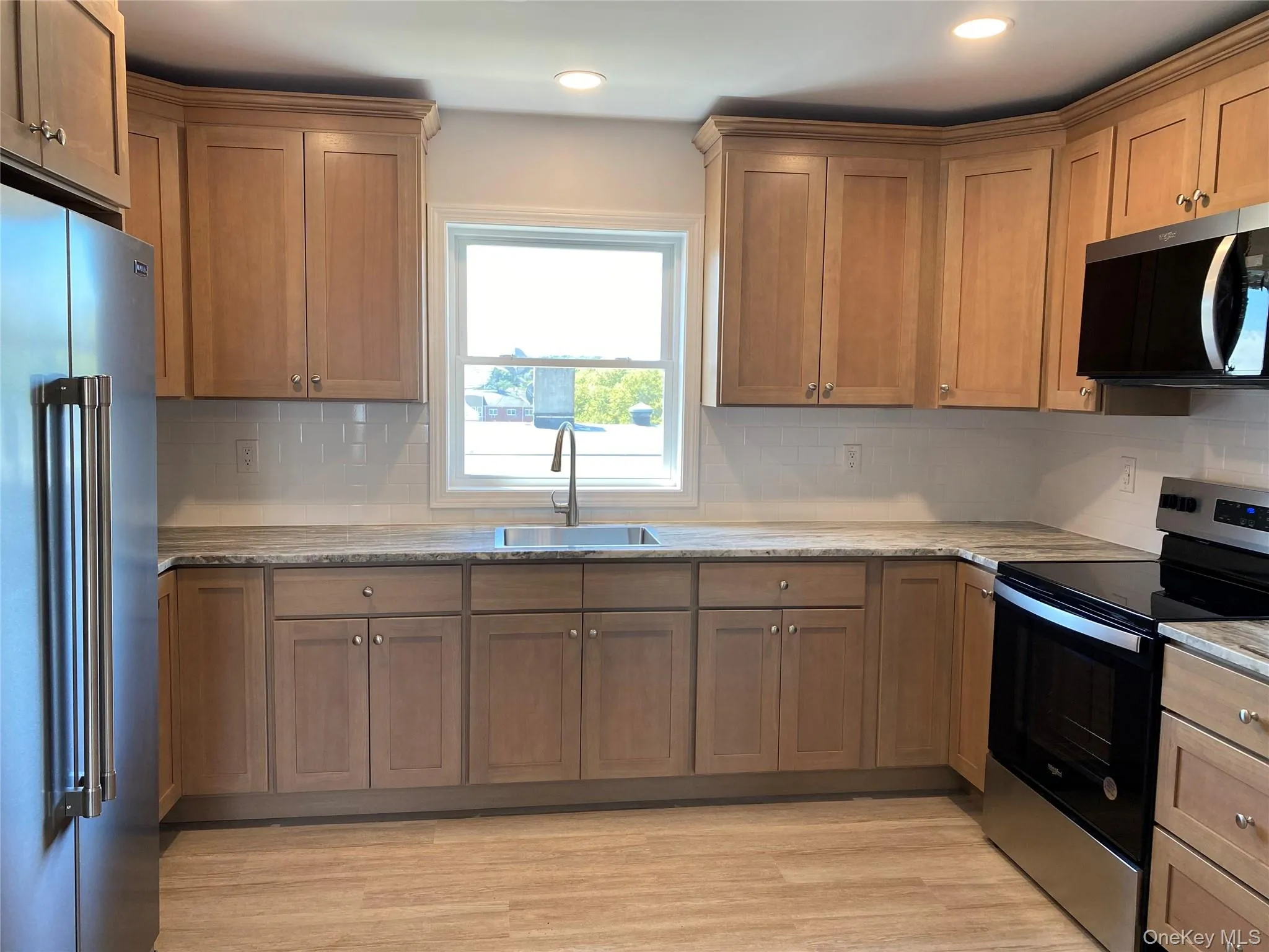 Kitchen with appliances with stainless steel finishes, decorative backsplash, light wood-type flooring, brown cabinets, and light stone counters Kitchen with appliances with stainless steel finishes, decorative backsplash, light wood-type flooring, brown cabinets, and light stone counters