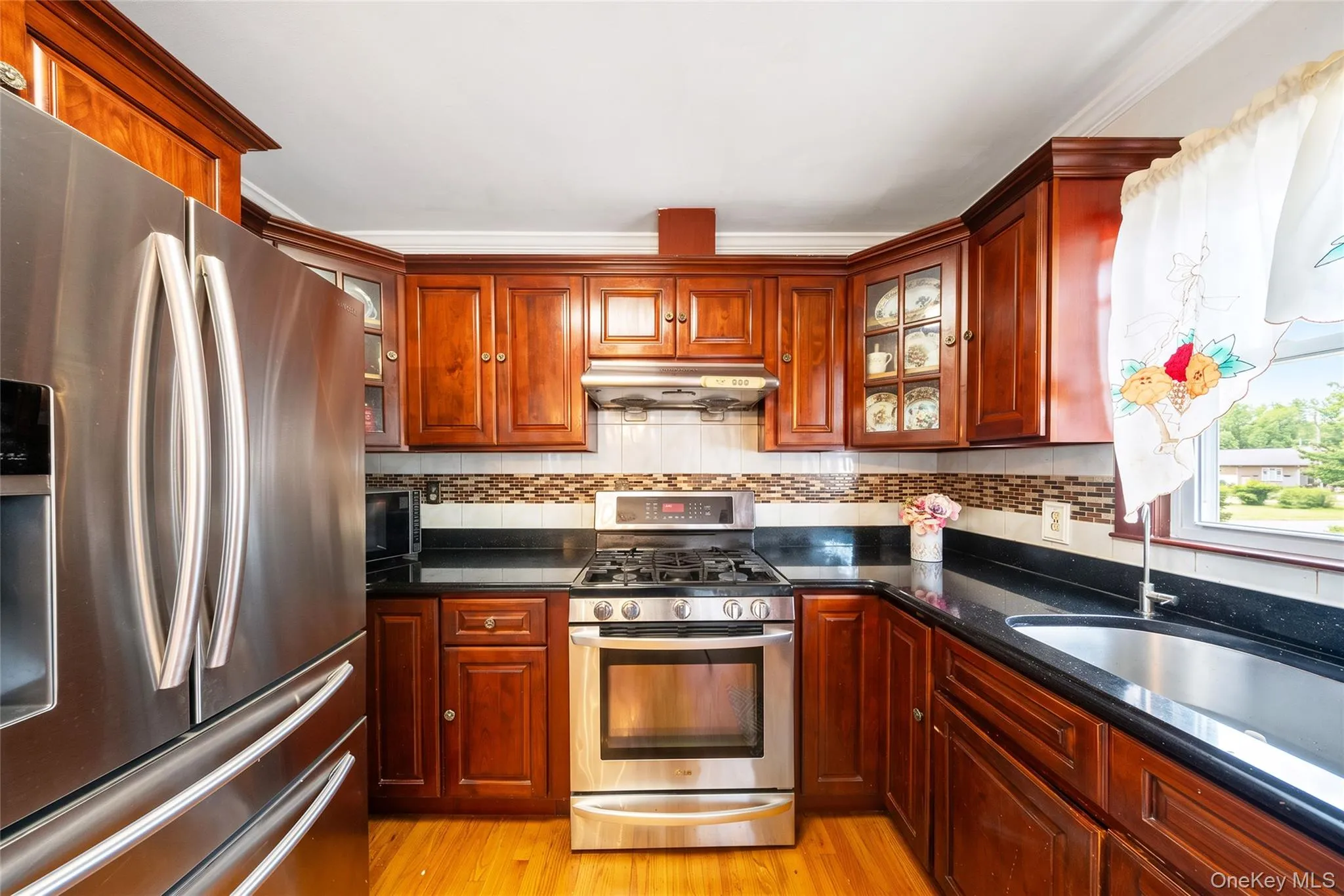 Wood-finish cabinetry with glass-front accents, dark countertops, stainless steel appliances, and wood-finish flooring Wood-finish cabinetry with glass-front accents, dark countertops, stainless steel appliances, and wood-finish flooring
