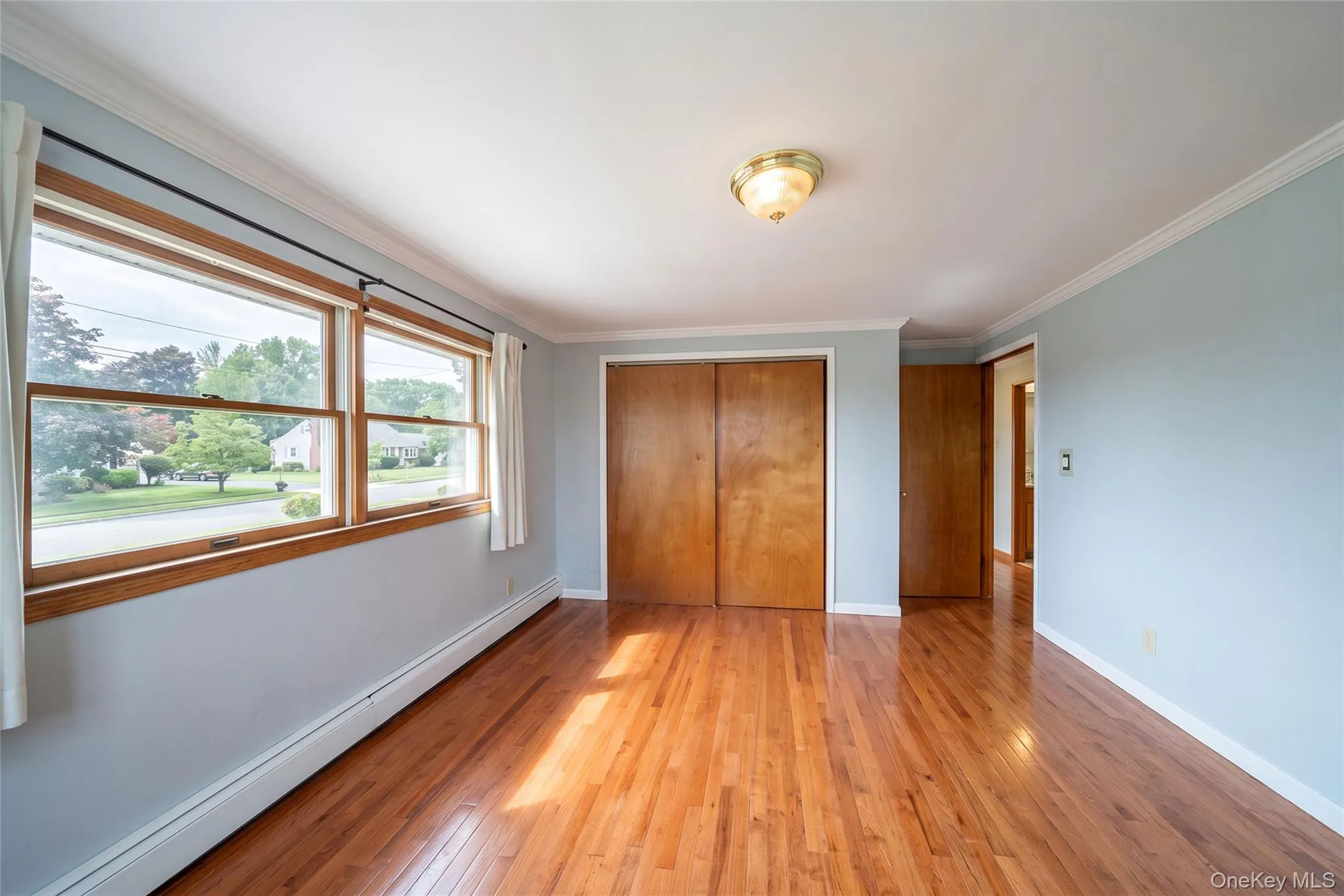 Hardwood flooring, two double-hung windows with wood trim, crown molding, double closet with bi-fold doors, and a flush mount ceiling light fixture Hardwood flooring, two double-hung windows with wood trim, crown molding, double closet with bi-fold doors, and a flush mount ceiling light fixture
