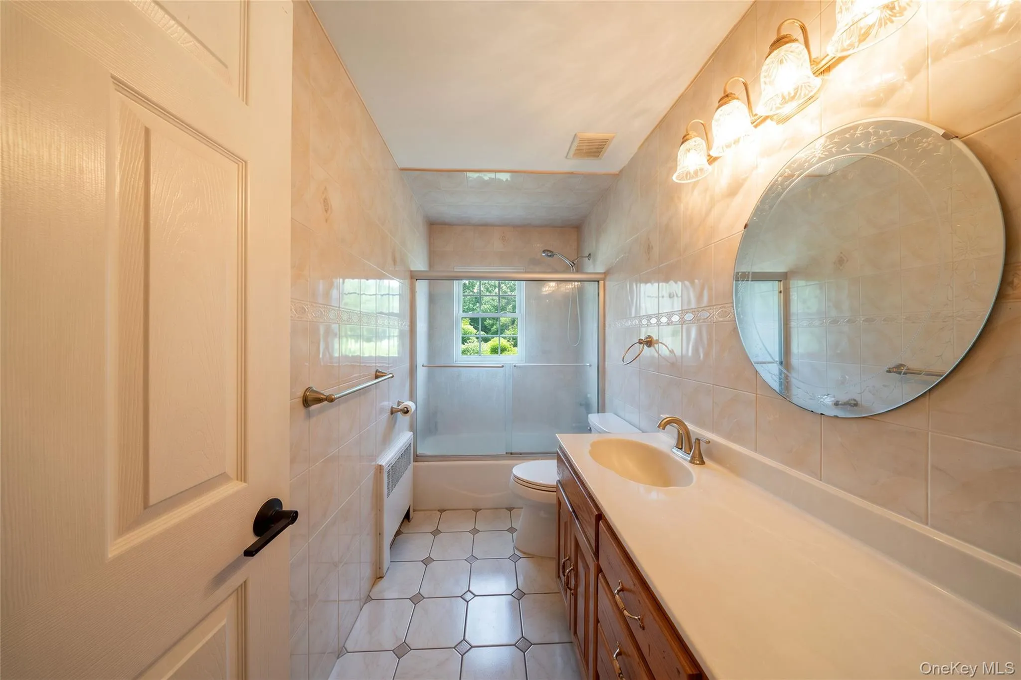 Ceramic tile flooring, full wall tiling, vanity with integrated sink, overhead lighting fixture, and a shower-tub combination with a glass enclosure Ceramic tile flooring, full wall tiling, vanity with integrated sink, overhead lighting fixture, and a shower-tub combination with a glass enclosure