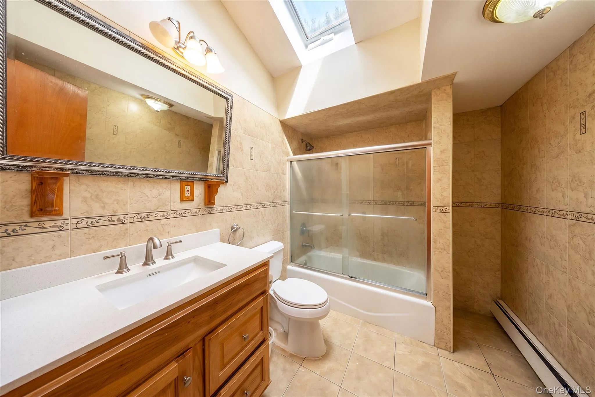 Tiled bathroom featuring a wood-finish vanity with a white countertop, a built-in bathtub with glass sliding doors, and a skylight Tiled bathroom featuring a wood-finish vanity with a white countertop, a built-in bathtub with glass sliding doors, and a skylight