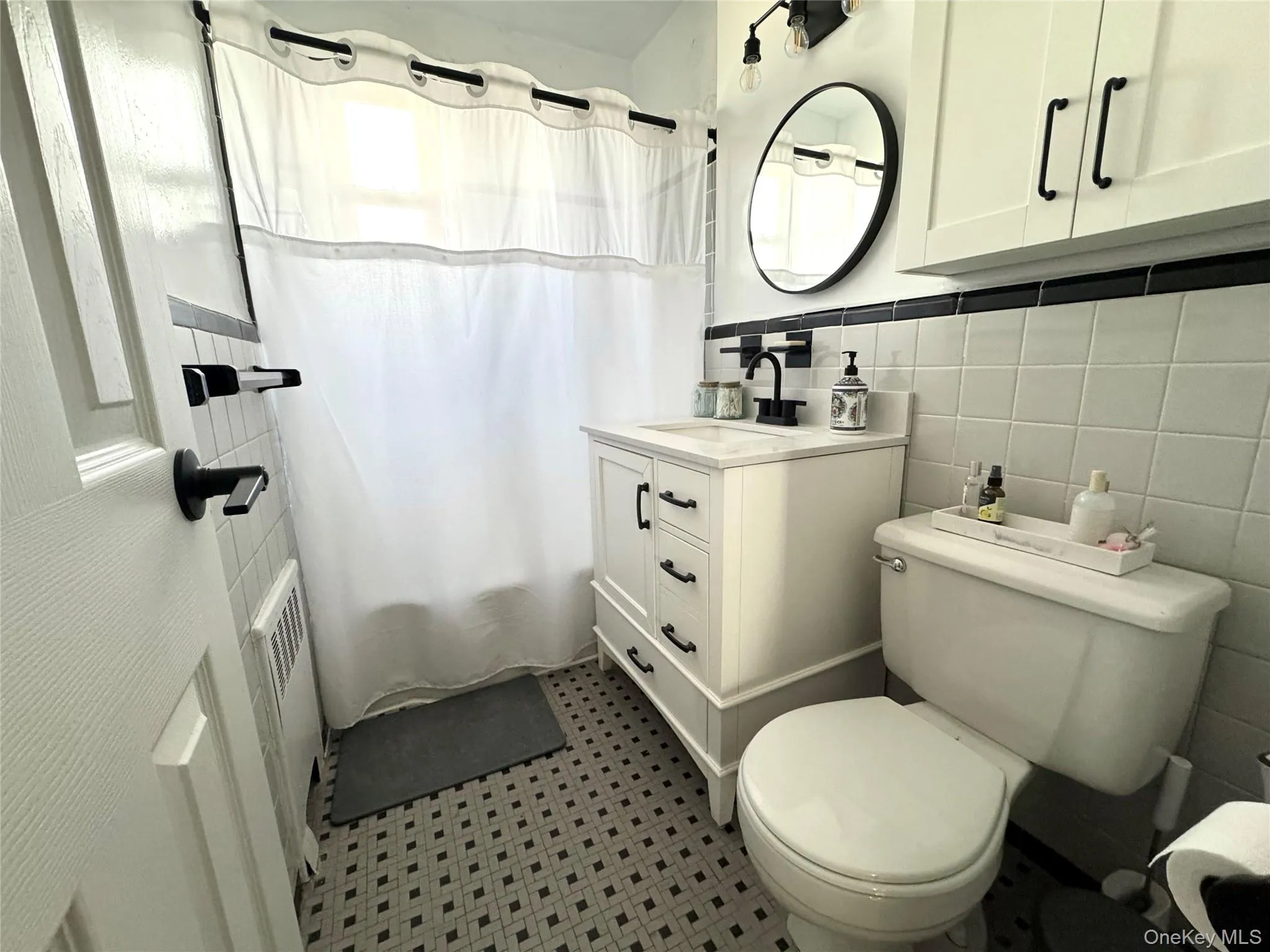 Vanity with integrated sink and vanity lights above circular mirror, black and gray tiled wall and flooring, wall-mounted cabinetry, black hardware accents, shower/tub combo with shower curtain Vanity with integrated sink and vanity lights above circular mirror, black and gray tiled wall and flooring, wall-mounted cabinetry, black hardware accents, shower/tub combo with shower curtain