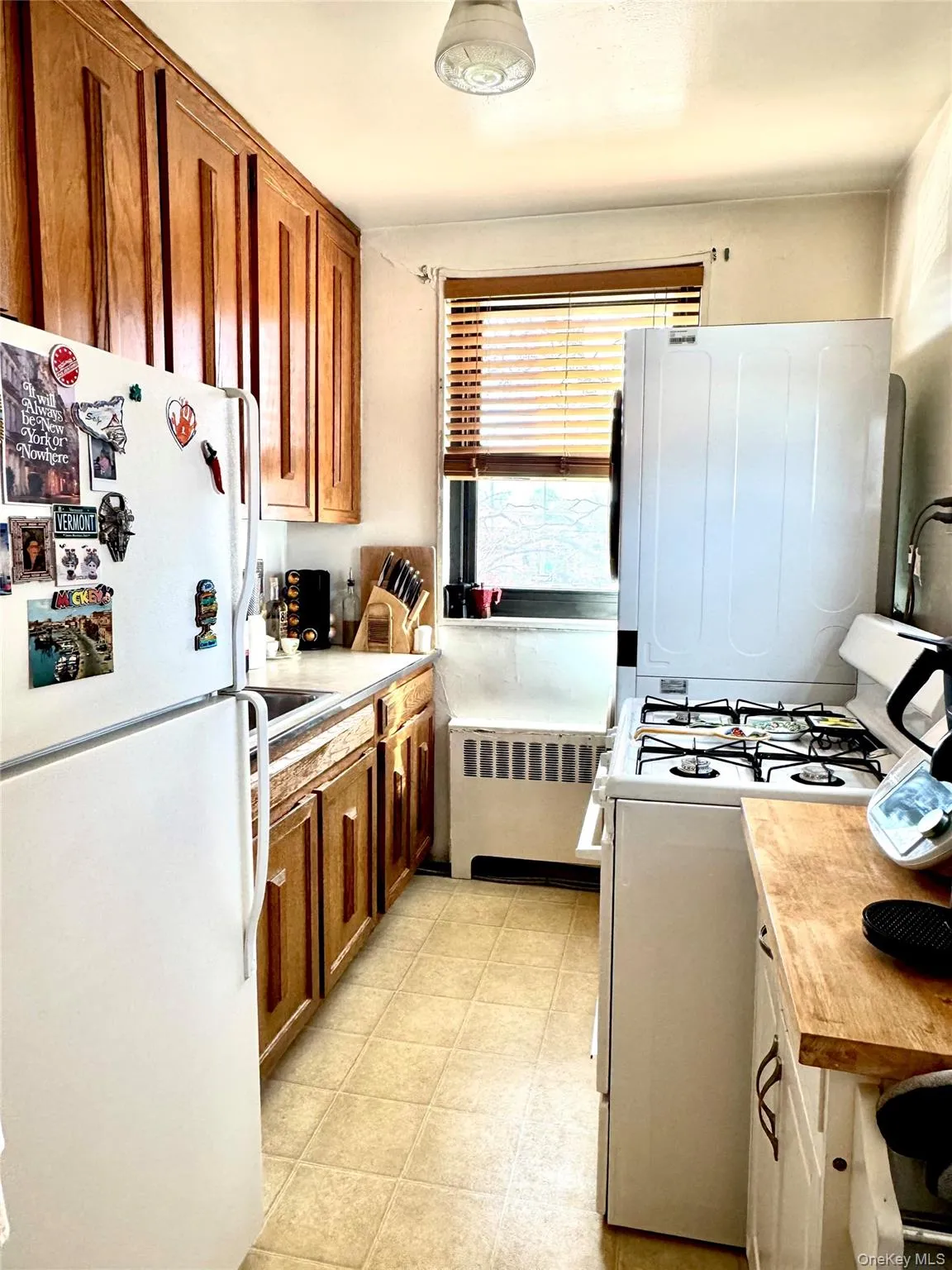 Galley kitchen featuring wood cabinetry, a window with blinds, tile flooring, white refrigerator, white gas stove and white stackable washer/dryer Galley kitchen featuring wood cabinetry, a window with blinds, tile flooring, white refrigerator, white gas stove and white stackable washer/dryer