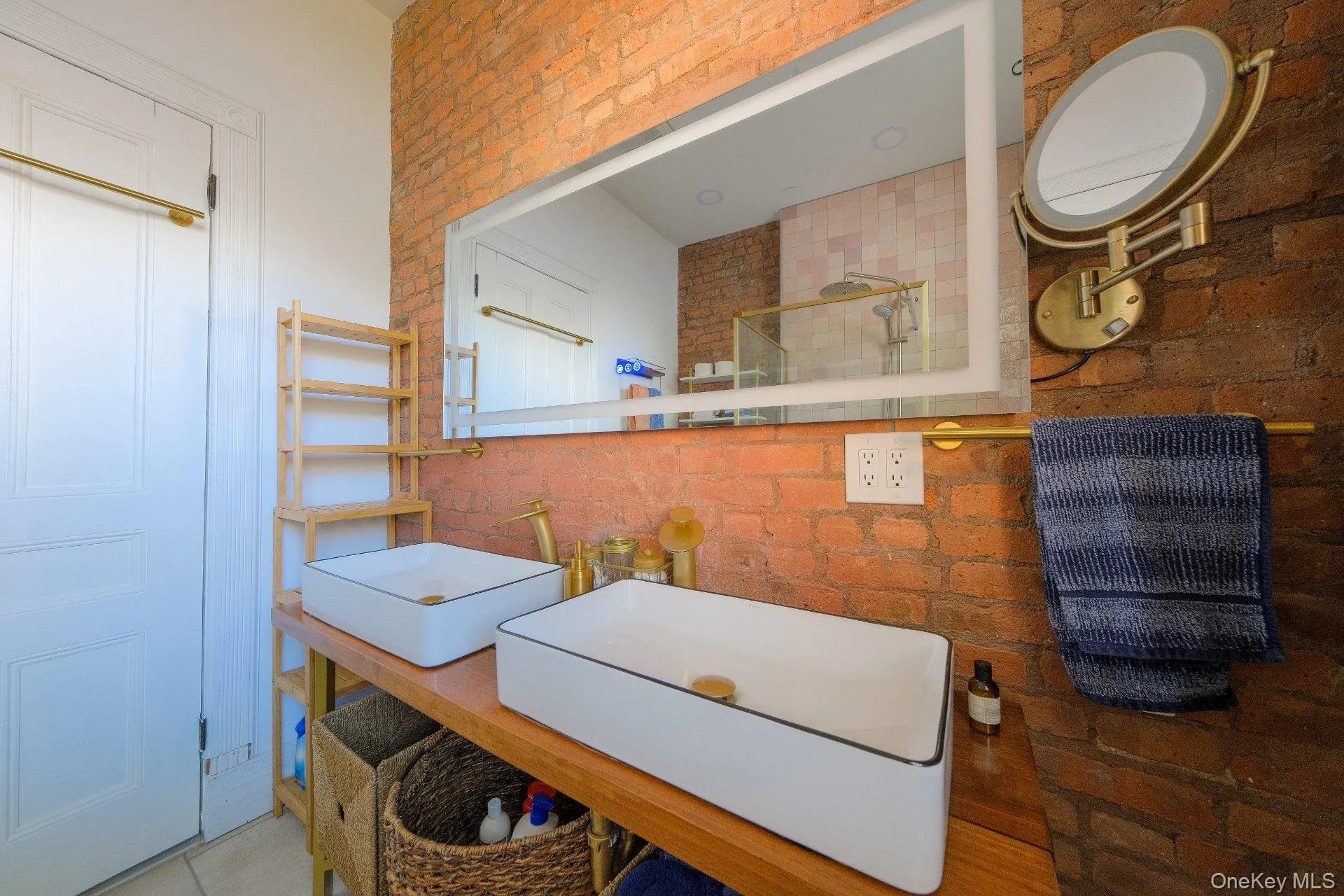 Exposed brick wall with a large back-lit mirror, dual vessel sinks, and wood-finish vanity Exposed brick wall with a large back-lit mirror, dual vessel sinks, and wood-finish vanity