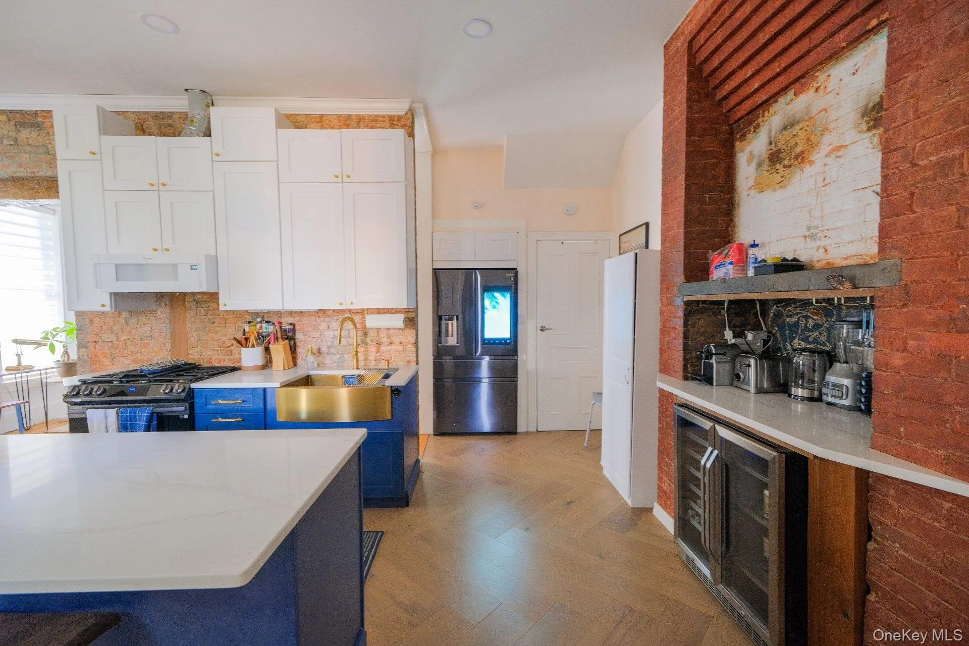 Kitchen featuring white upper cabinetry, blue lower cabinetry, a gold-tone sink, a herringbone wood-finish floor, and exposed brick accents Kitchen featuring white upper cabinetry, blue lower cabinetry, a gold-tone sink, a herringbone wood-finish floor, and exposed brick accents