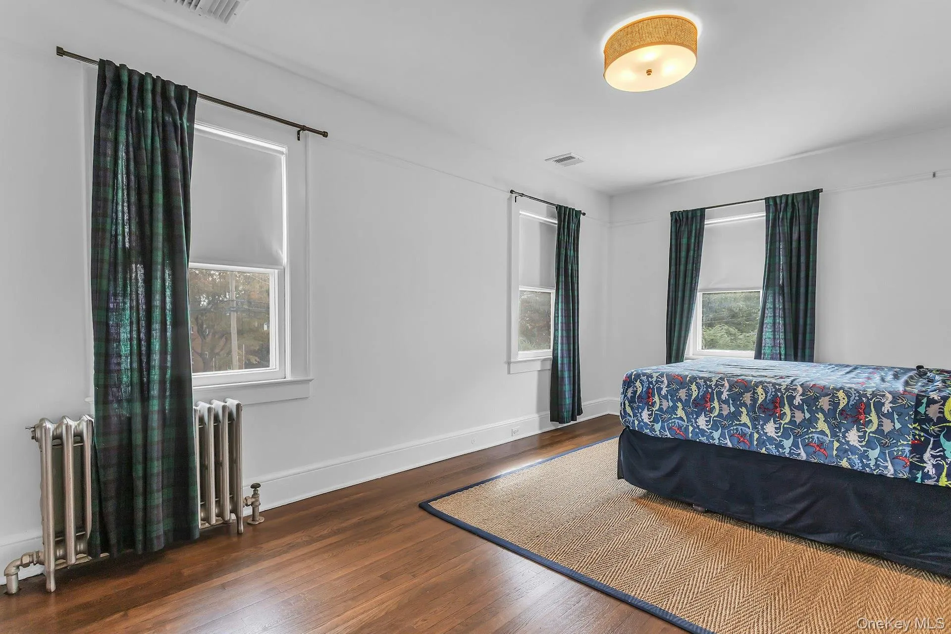 Hardwood flooring throughout, multiple windows with roller shades, cast iron radiator, and ceiling-mounted light fixture Hardwood flooring throughout, multiple windows with roller shades, cast iron radiator, and ceiling-mounted light fixture