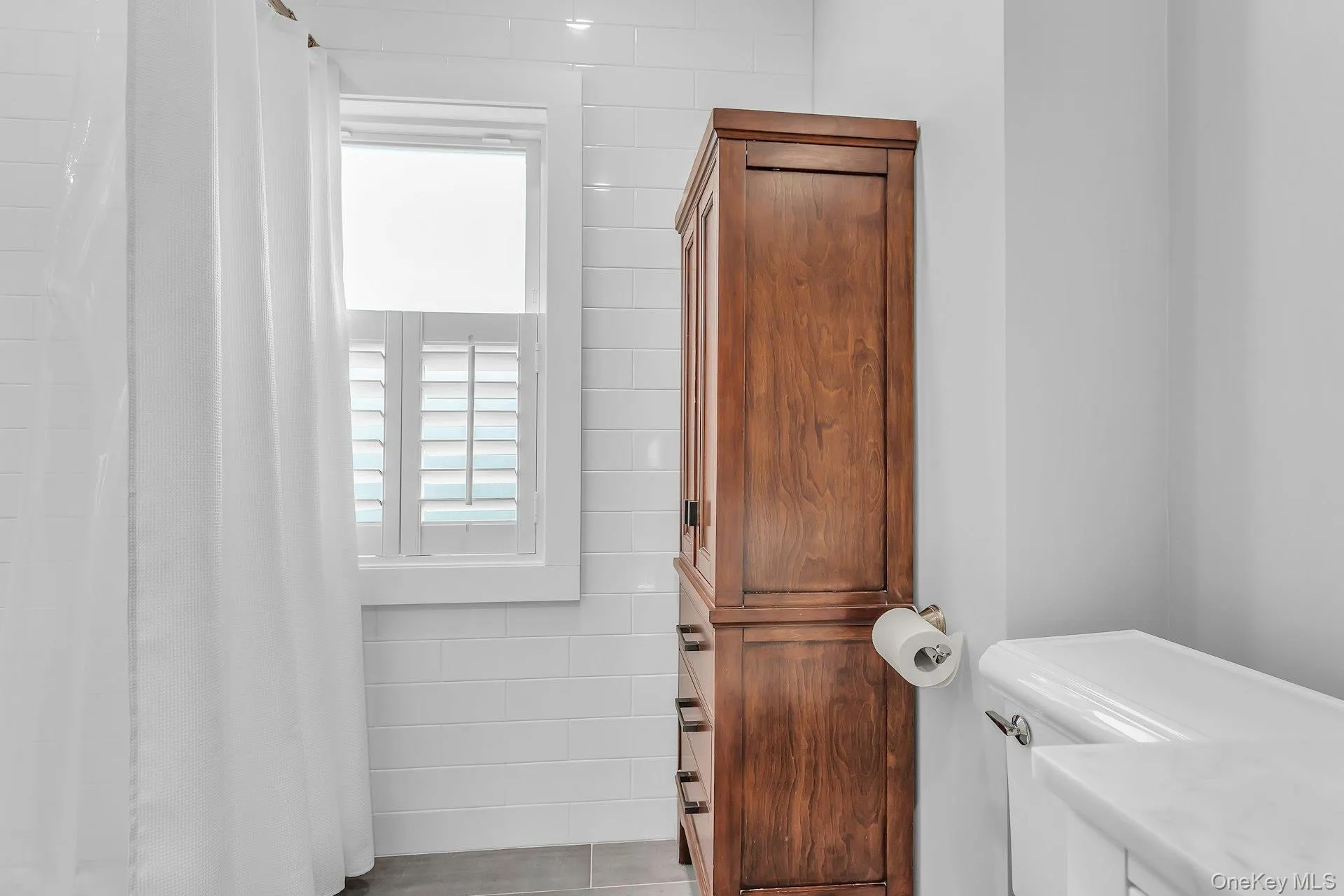 White subway tile walls, wood-finish storage cabinet, window with plantation shutters, white toilet, and gray floor tiles White subway tile walls, wood-finish storage cabinet, window with plantation shutters, white toilet, and gray floor tiles