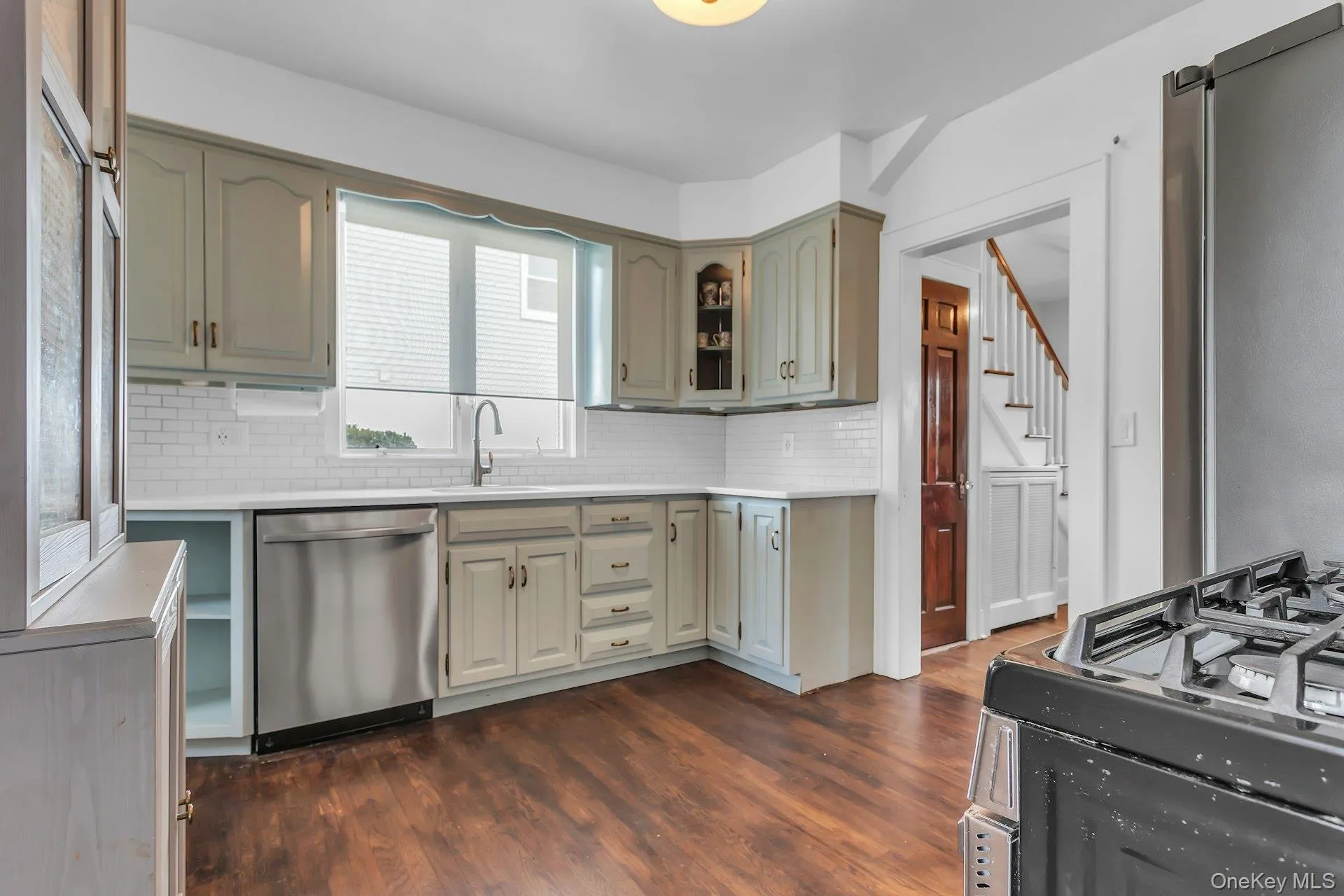 Kitchen featuring wood-finish flooring, white subway tile backsplash, light-colored cabinetry, stainless steel dishwasher, and a window above the sink Kitchen featuring wood-finish flooring, white subway tile backsplash, light-colored cabinetry, stainless steel dishwasher, and a window above the sink