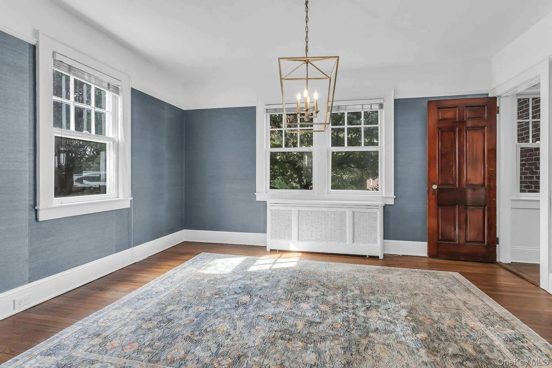 Hardwood flooring with a matte finish, blue textured wall covering, a dark wood panel door, white window casings, and a geometric chandelier Hardwood flooring with a matte finish, blue textured wall covering, a dark wood panel door, white window casings, and a geometric chandelier