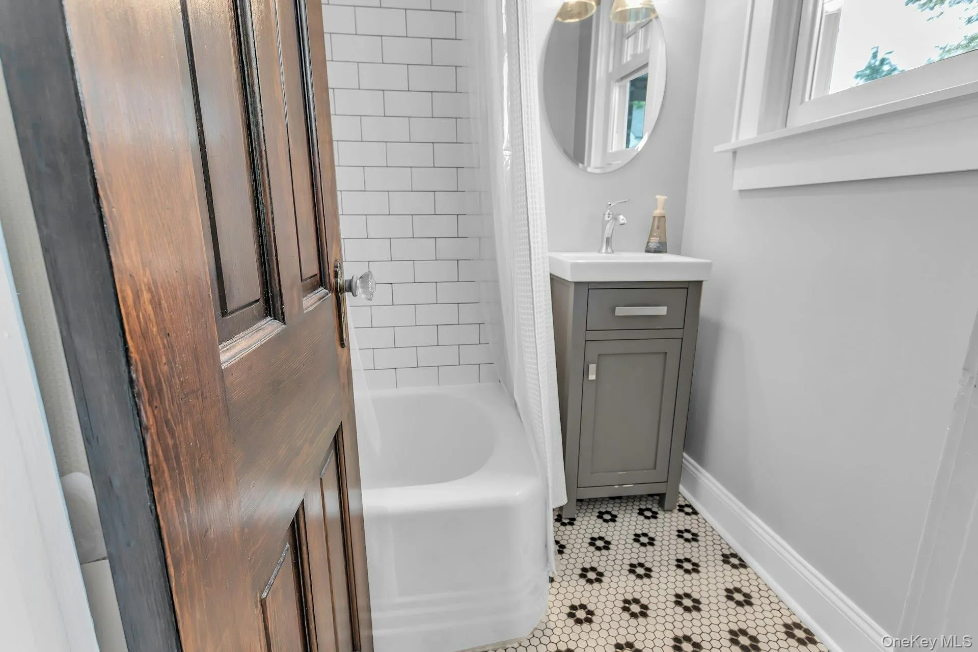 Subway tile bath surround, white tub, gray vanity with integrated sink, patterned mosaic floor tile, and an oval mirror Subway tile bath surround, white tub, gray vanity with integrated sink, patterned mosaic floor tile, and an oval mirror