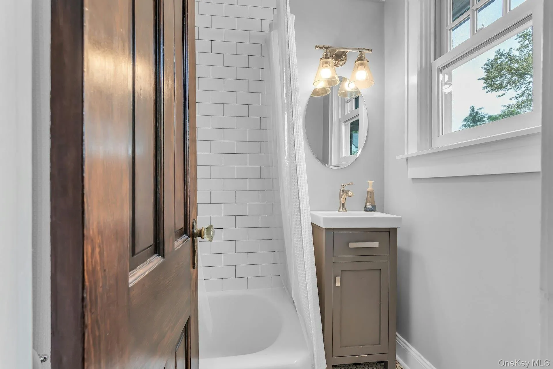 White subway tile shower surround, single window with white trim, gray vanity with integrated sink, gold-toned faucet, and oval wall mirror White subway tile shower surround, single window with white trim, gray vanity with integrated sink, gold-toned faucet, and oval wall mirror