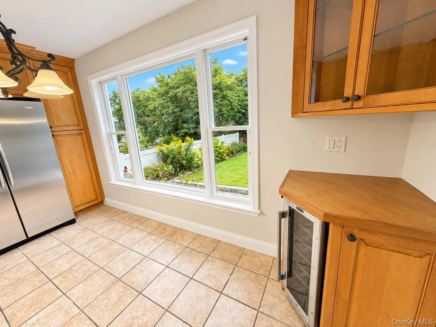 Tile flooring, light wood cabinetry, integrated beverage cooler, and a large window overlooking a landscaped yard Tile flooring, light wood cabinetry, integrated beverage cooler, and a large window overlooking a landscaped yard