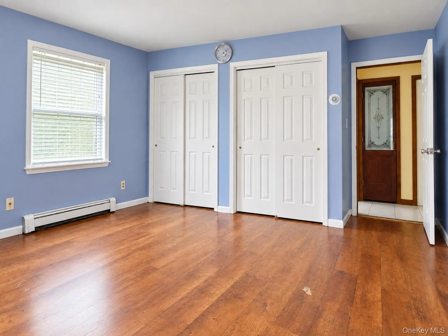 Wood-finish flooring with white baseboards, two sets of white paneled bifold closet doors, and a single window with blinds Wood-finish flooring with white baseboards, two sets of white paneled bifold closet doors, and a single window with blinds