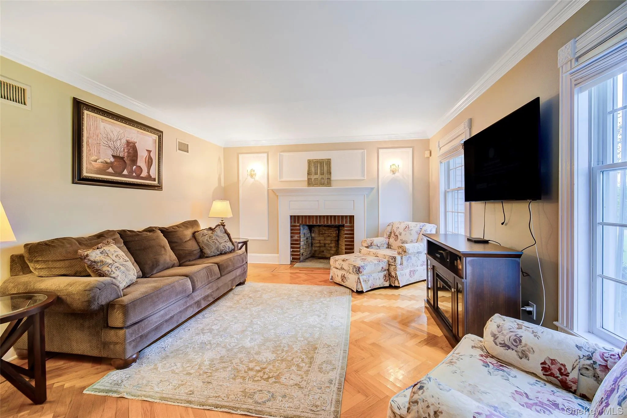 Living room area featuring herringbone wood flooring, crown molding, a brick fireplace with a white mantel, and large windows with white trim Living room area featuring herringbone wood flooring, crown molding, a brick fireplace with a white mantel, and large windows with white trim