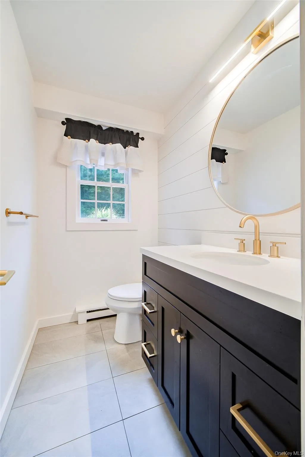 First floor 1/2 bath features wood-finish vanity with gold-tone hardware, white stone countertop, round gold-tone framed mirror, tiled flooring and white shiplap-style accent wall,. First floor 1/2 bath features wood-finish vanity with gold-tone hardware, white stone countertop, round gold-tone framed mirror, tiled flooring and white shiplap-style accent wall,.
