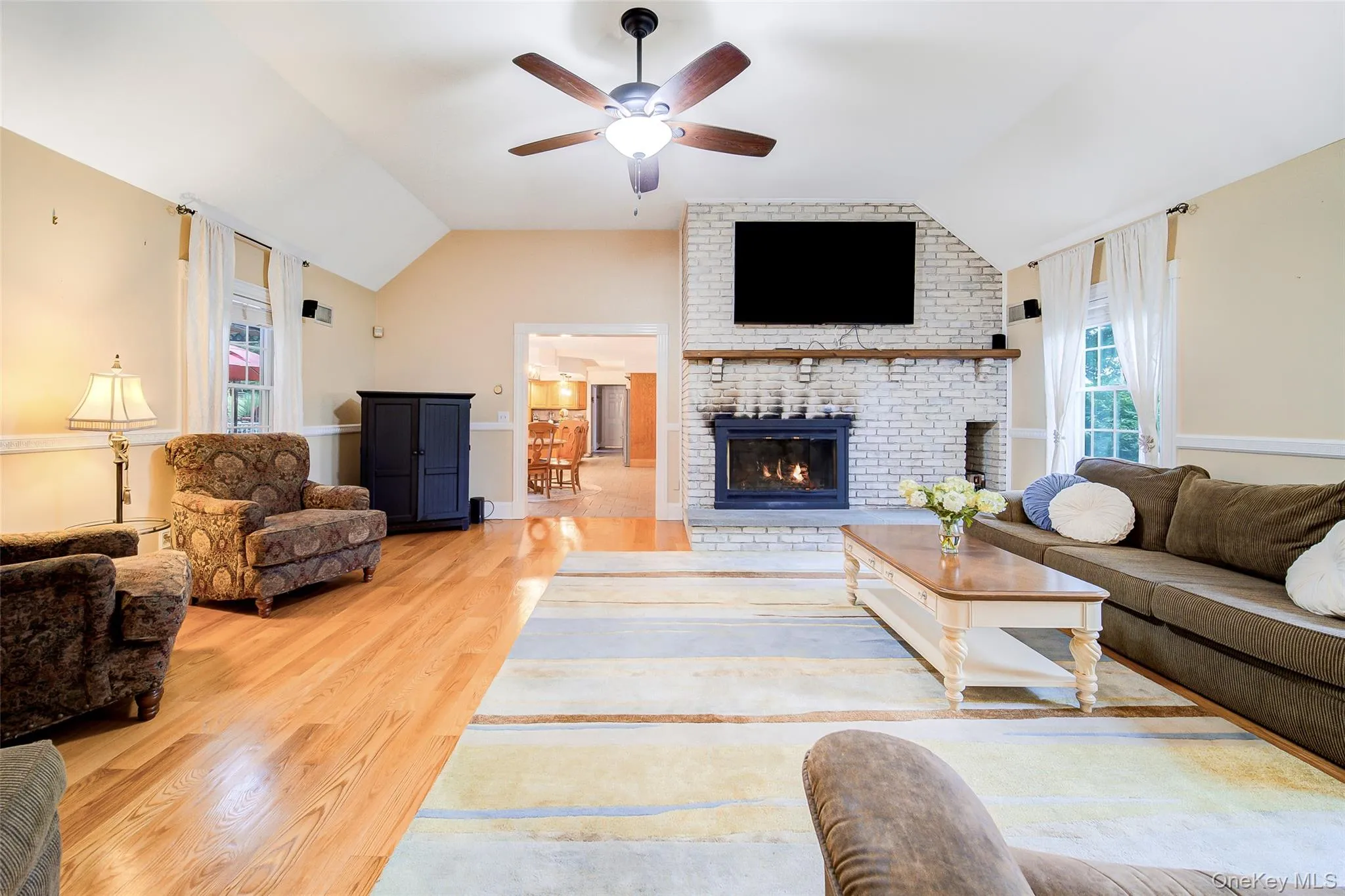 Vaulted ceiling, ceiling fan with integrated lighting, natural wood-finish flooring, white-painted brick fireplace with wood mantel, and multiple windows for natural lighting Vaulted ceiling, ceiling fan with integrated lighting, natural wood-finish flooring, white-painted brick fireplace with wood mantel, and multiple windows for natural lighting