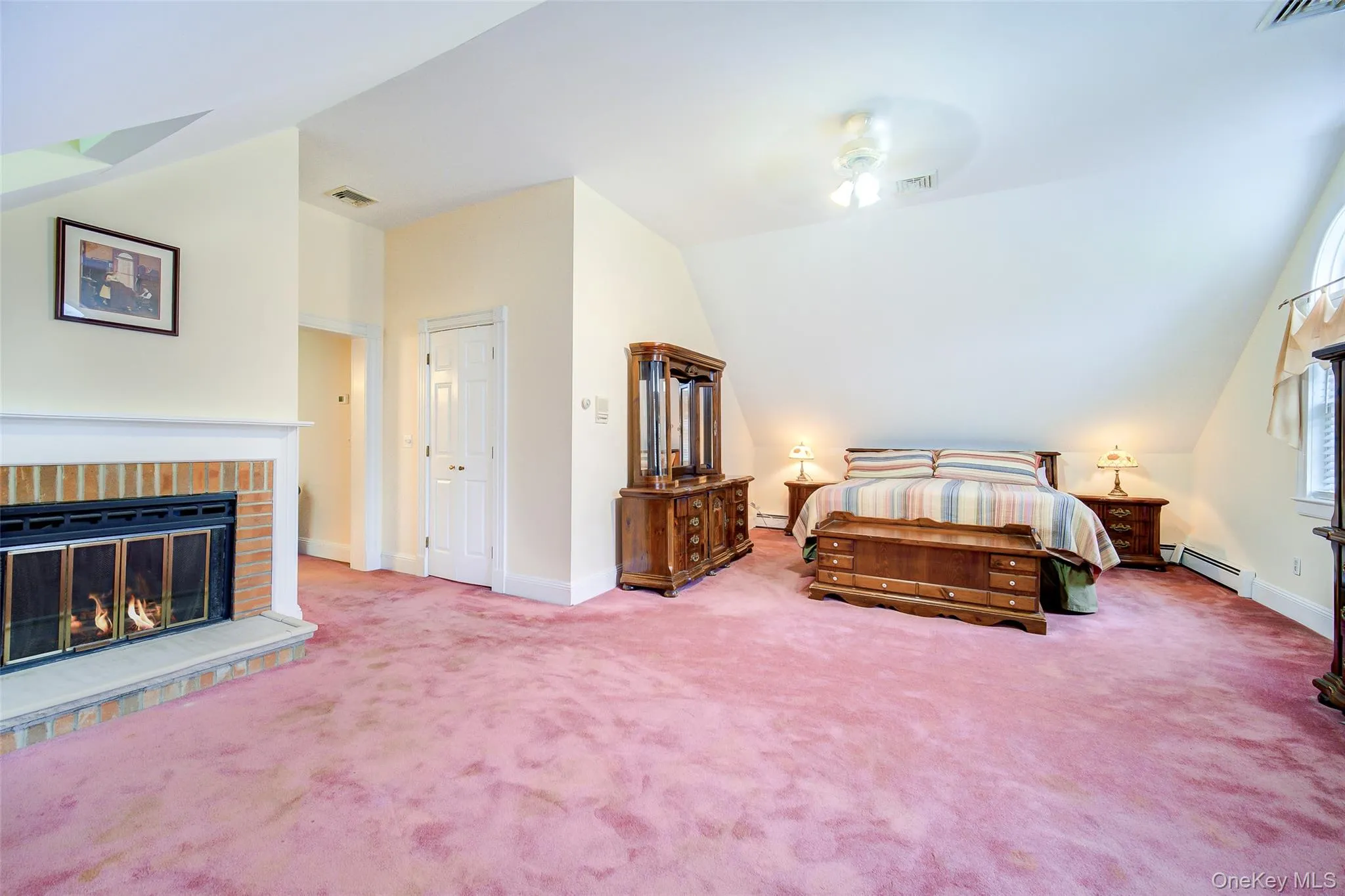 This spacious room features a brick fireplace with a mantel, sloped ceilings, and and arched window. This spacious room features a brick fireplace with a mantel, sloped ceilings, and and arched window.