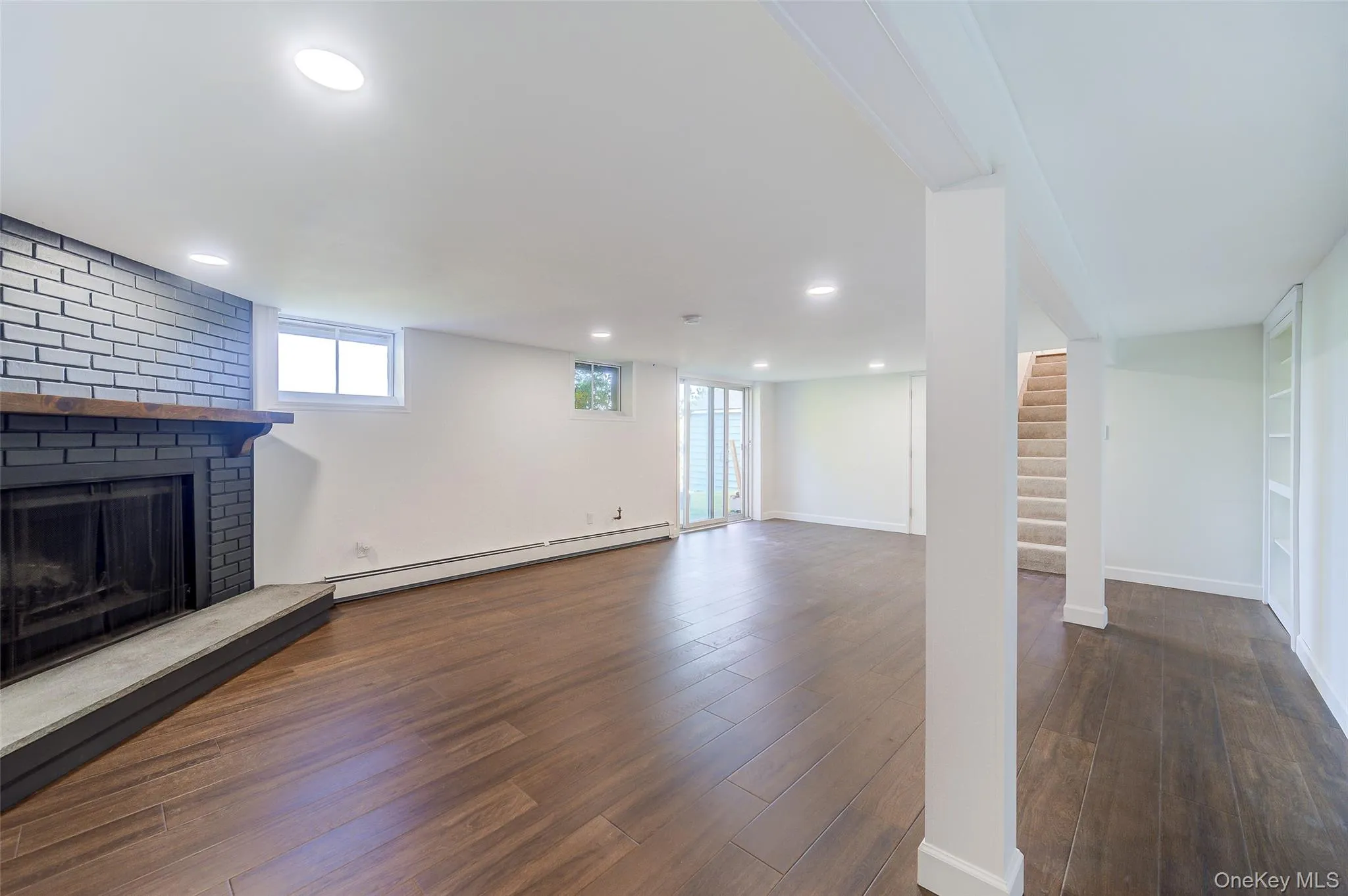 Spacious room featuring wood-finish flooring, recessed lighting, and a decorative fireplace with a black brick surround and stone hearth Spacious room featuring wood-finish flooring, recessed lighting, and a decorative fireplace with a black brick surround and stone hearth