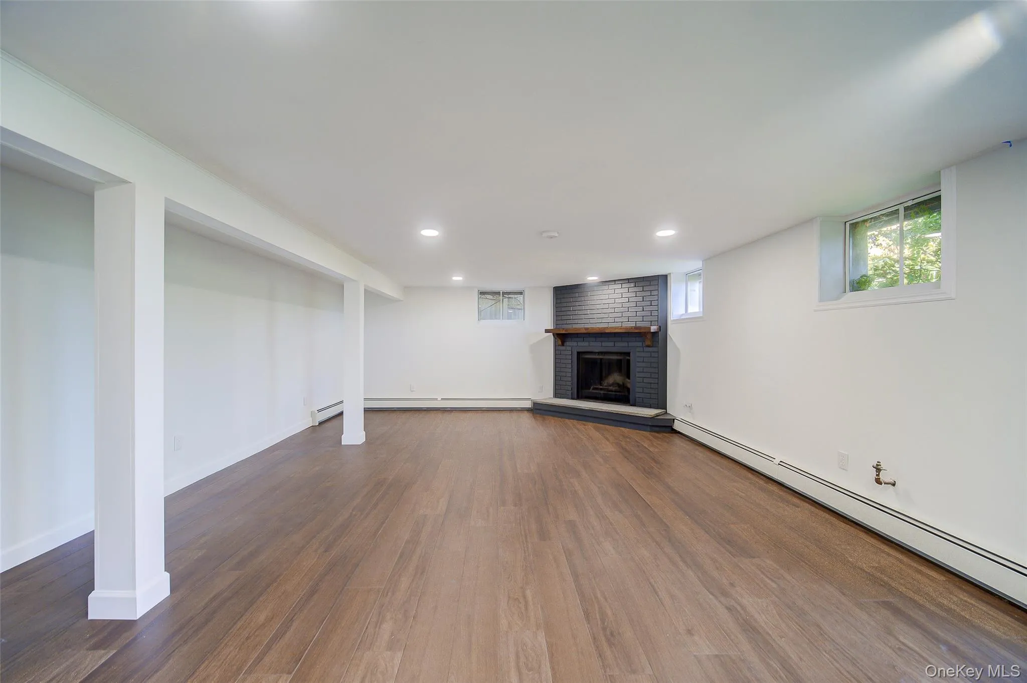 Wood-finish flooring, recessed lighting, white walls, black brick-surround fireplace (decorative) with custom wood mantel, and multiple windows Wood-finish flooring, recessed lighting, white walls, black brick-surround fireplace (decorative) with custom wood mantel, and multiple windows