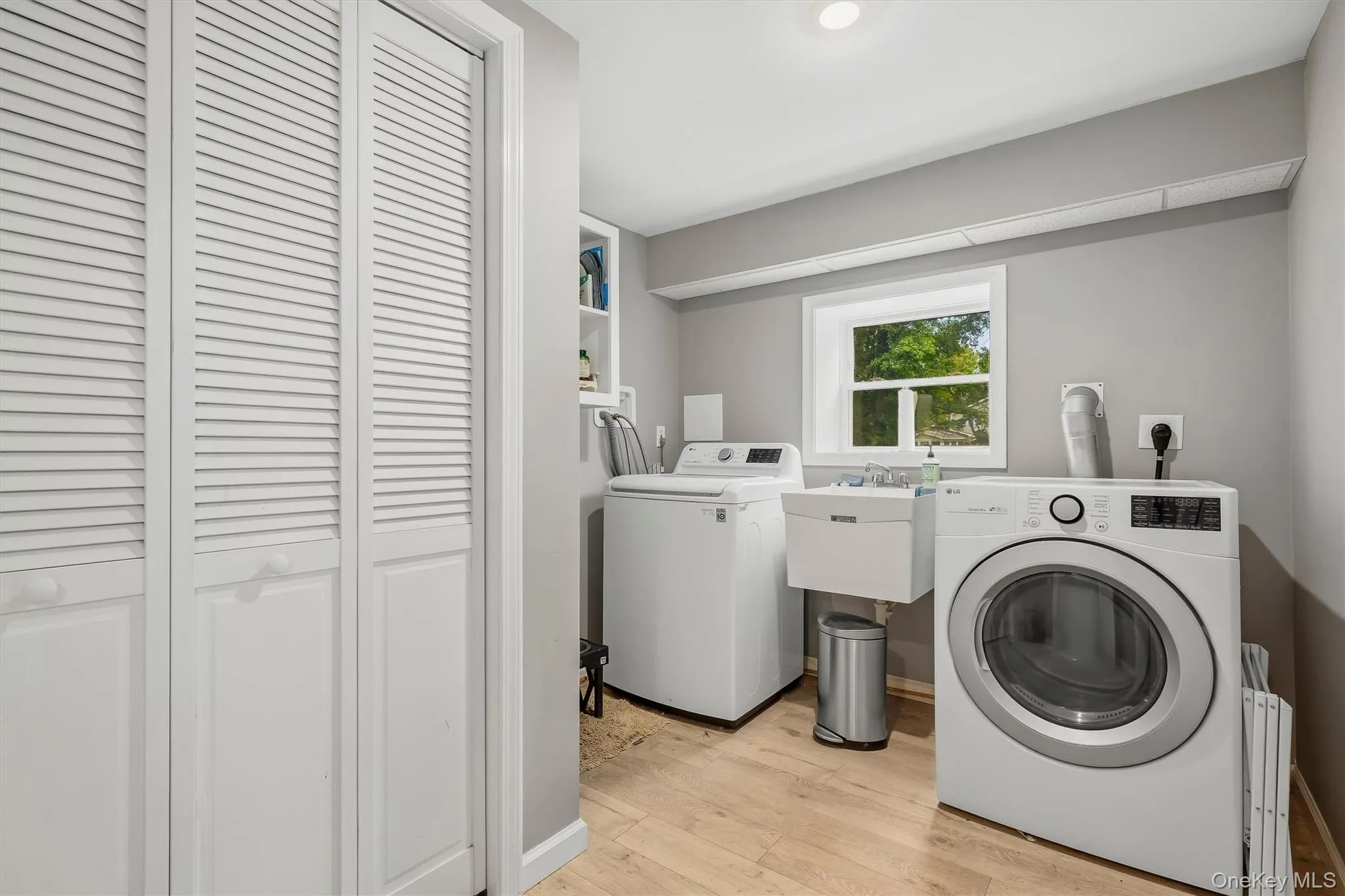 White louvered bifold doors, window with natural light, utility sink, overhead lighting, and wood-finish flooring White louvered bifold doors, window with natural light, utility sink, overhead lighting, and wood-finish flooring