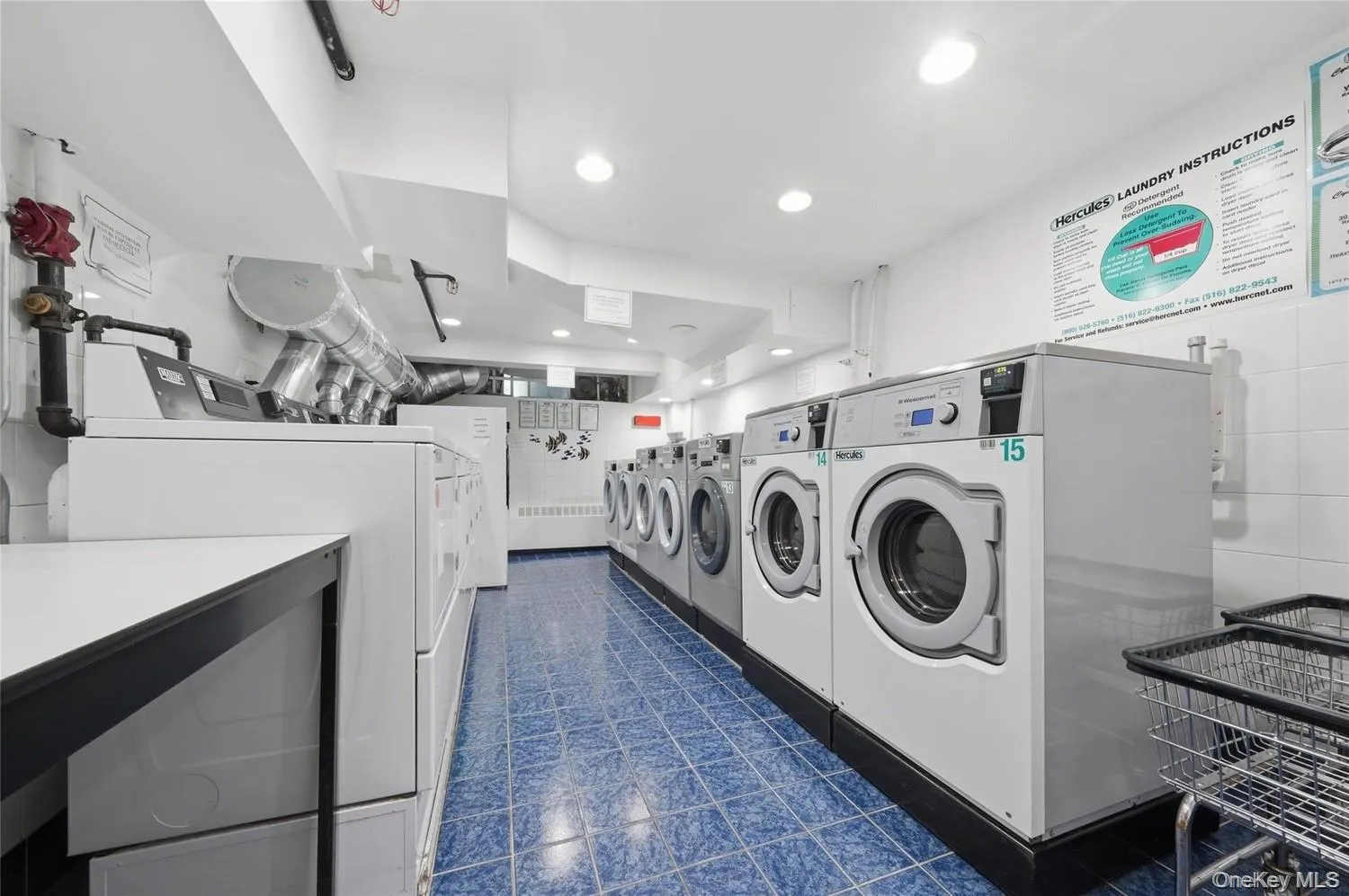 Professional laundry facility featuring multiple front-load washing machines and dryers, stainless steel appliances, blue-toned polished concrete flooring, recessed ceiling lighting, and white cabinetry with overhead storage Professional laundry facility featuring multiple front-load washing machines and dryers, stainless steel appliances, blue-toned polished concrete flooring, recessed ceiling lighting, and white cabinetry with overhead storage