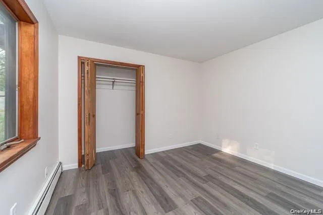 Wood-finish flooring with light-toned walls, featuring a window with wood trim and a bifold door closet with an integrated hanging rod Wood-finish flooring with light-toned walls, featuring a window with wood trim and a bifold door closet with an integrated hanging rod