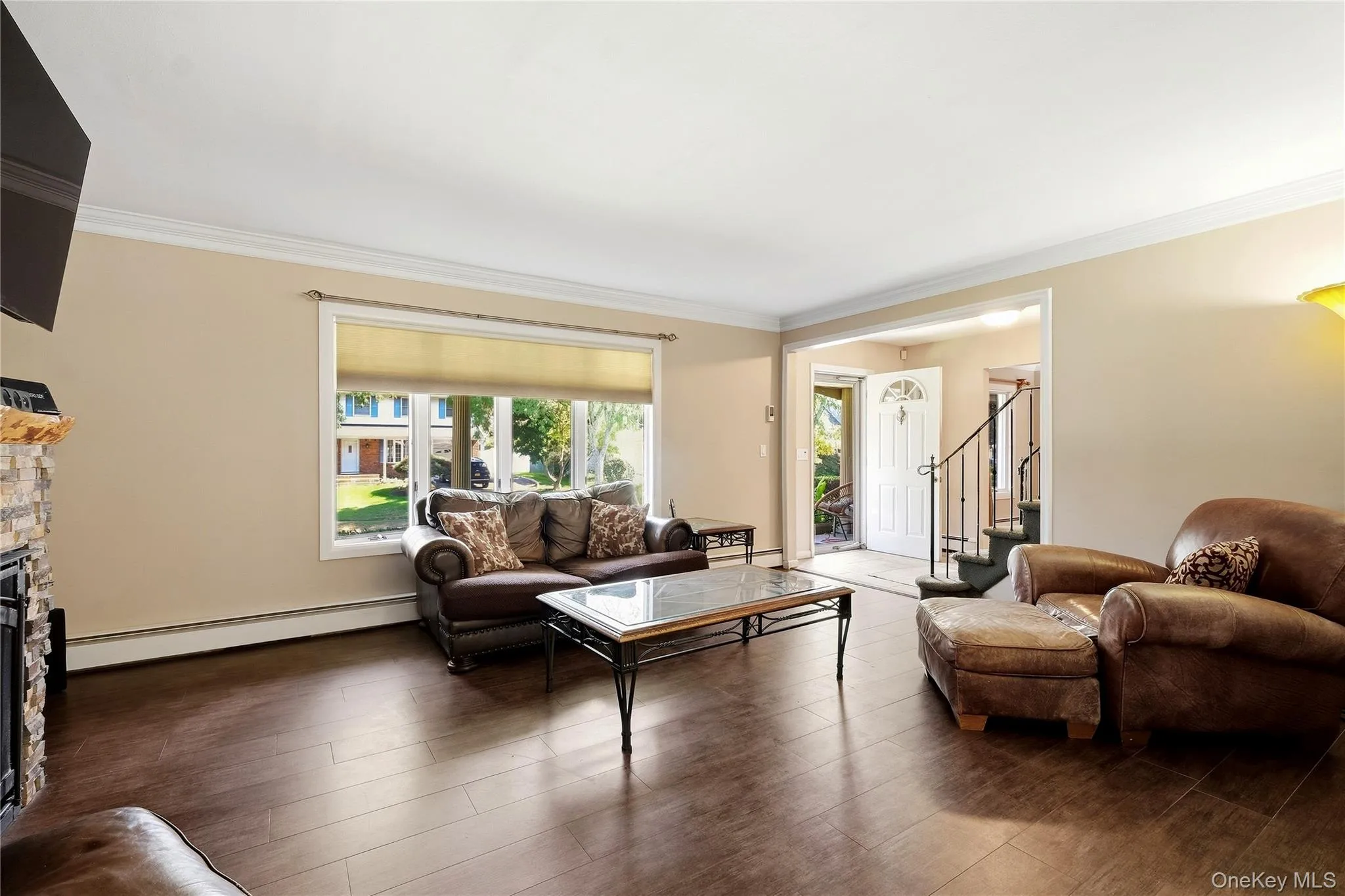 Wood-finish flooring, neutral wall tones, stone-clad fireplace with hearth, crown molding, and a large front-facing window Wood-finish flooring, neutral wall tones, stone-clad fireplace with hearth, crown molding, and a large front-facing window