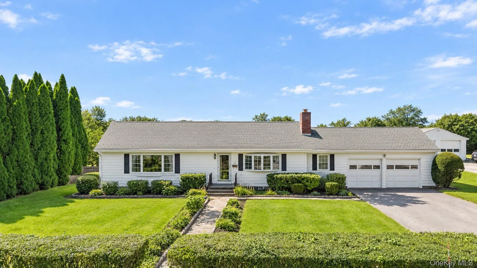 White ranch-style residence featuring a two-car garage, brick chimney, established front landscaping, and a paved driveway White ranch-style residence featuring a two-car garage, brick chimney, established front landscaping, and a paved driveway
