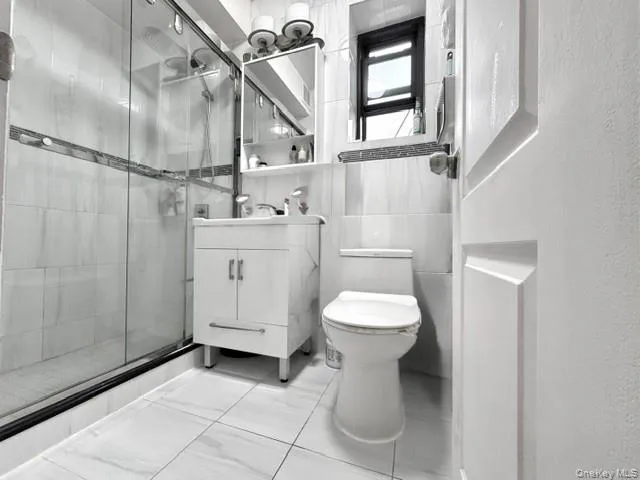 Shower with sliding glass enclosure, white tile flooring, white vanity with integrated sink, wall-mounted mirror cabinet, and a window Shower with sliding glass enclosure, white tile flooring, white vanity with integrated sink, wall-mounted mirror cabinet, and a window