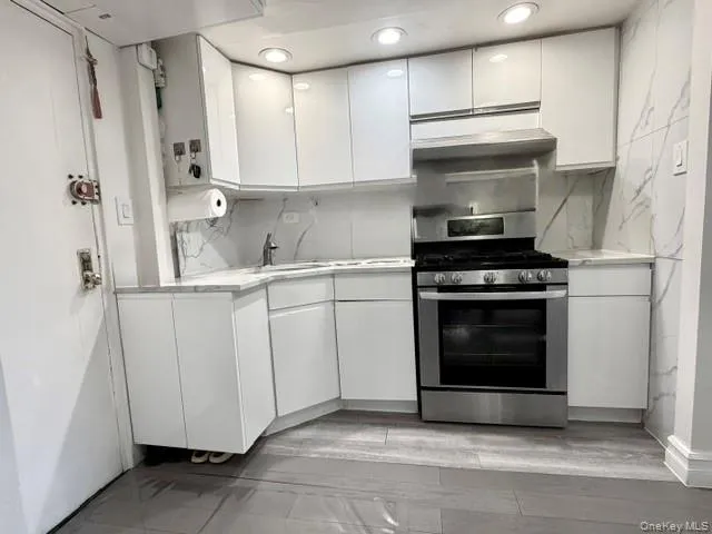 White gloss cabinetry, stainless steel gas range, integrated range hood, recessed lighting, and stone-finish countertops with matching backsplash White gloss cabinetry, stainless steel gas range, integrated range hood, recessed lighting, and stone-finish countertops with matching backsplash