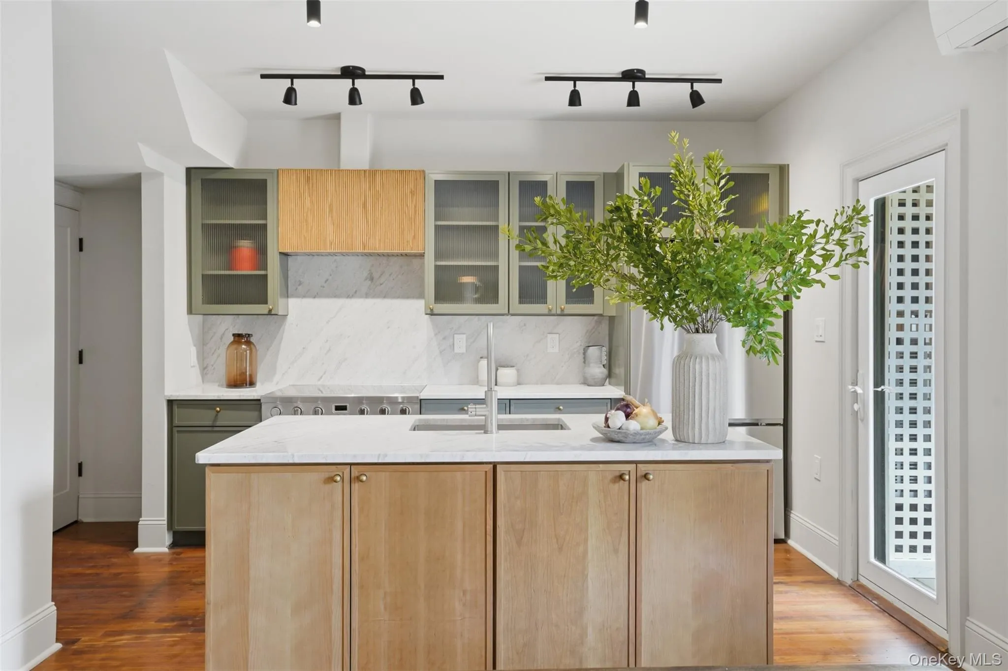 Kitchen featuring a central island with wood-finish cabinetry and a light-toned countertop, a range with a stainless steel cooktop, light-toned backsplash, and a combination of green and wood-finish upper cabinetry with glass inserts Kitchen featuring a central island with wood-finish cabinetry and a light-toned countertop, a range with a stainless steel cooktop, light-toned backsplash, and a combination of green and wood-finish upper cabinetry with glass inserts