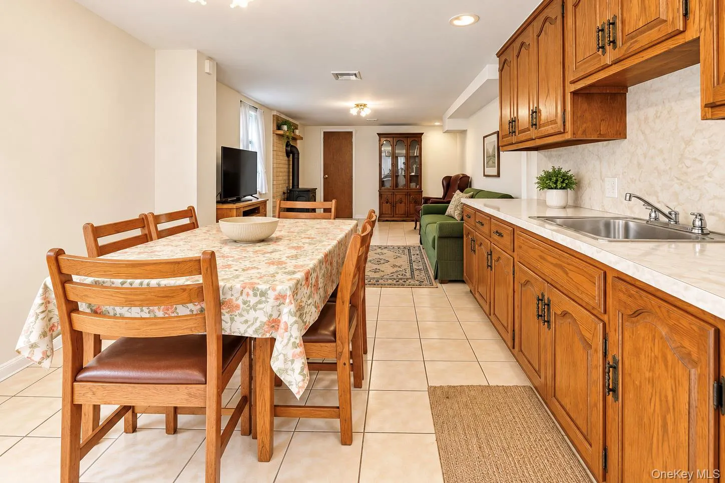 Extended kitchen and dining area featuring wood-finish cabinetry, a stainless steel sink, light-toned countertops, and tile flooring Extended kitchen and dining area featuring wood-finish cabinetry, a stainless steel sink, light-toned countertops, and tile flooring