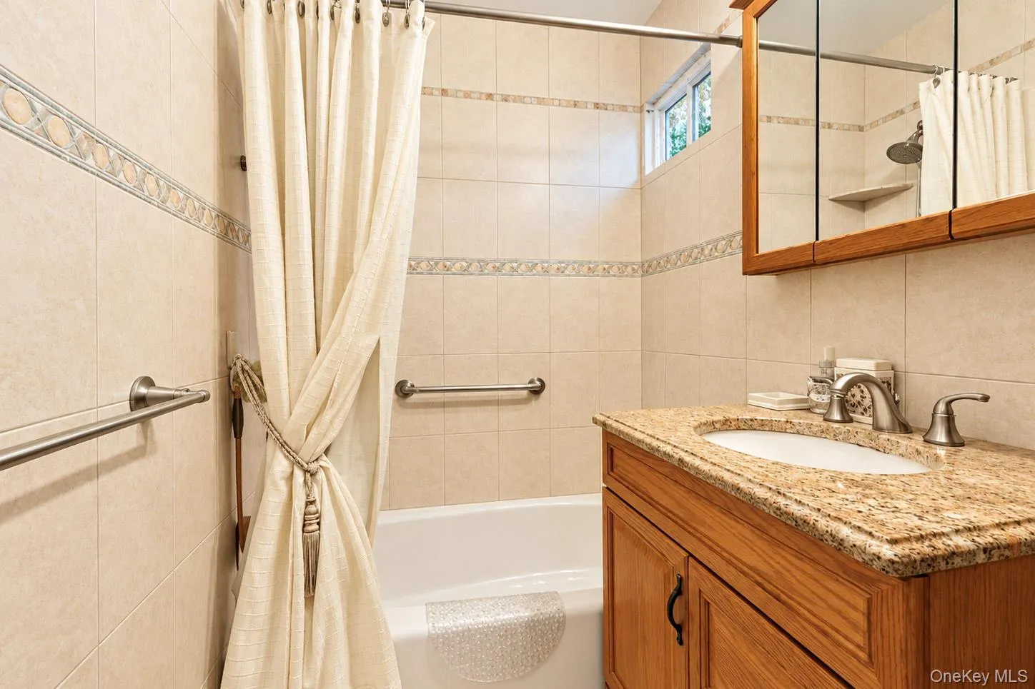 Tiled bathroom featuring a built-in bathtub with shower, wood-finish vanity, and a light-colored countertop with an integrated sink Tiled bathroom featuring a built-in bathtub with shower, wood-finish vanity, and a light-colored countertop with an integrated sink